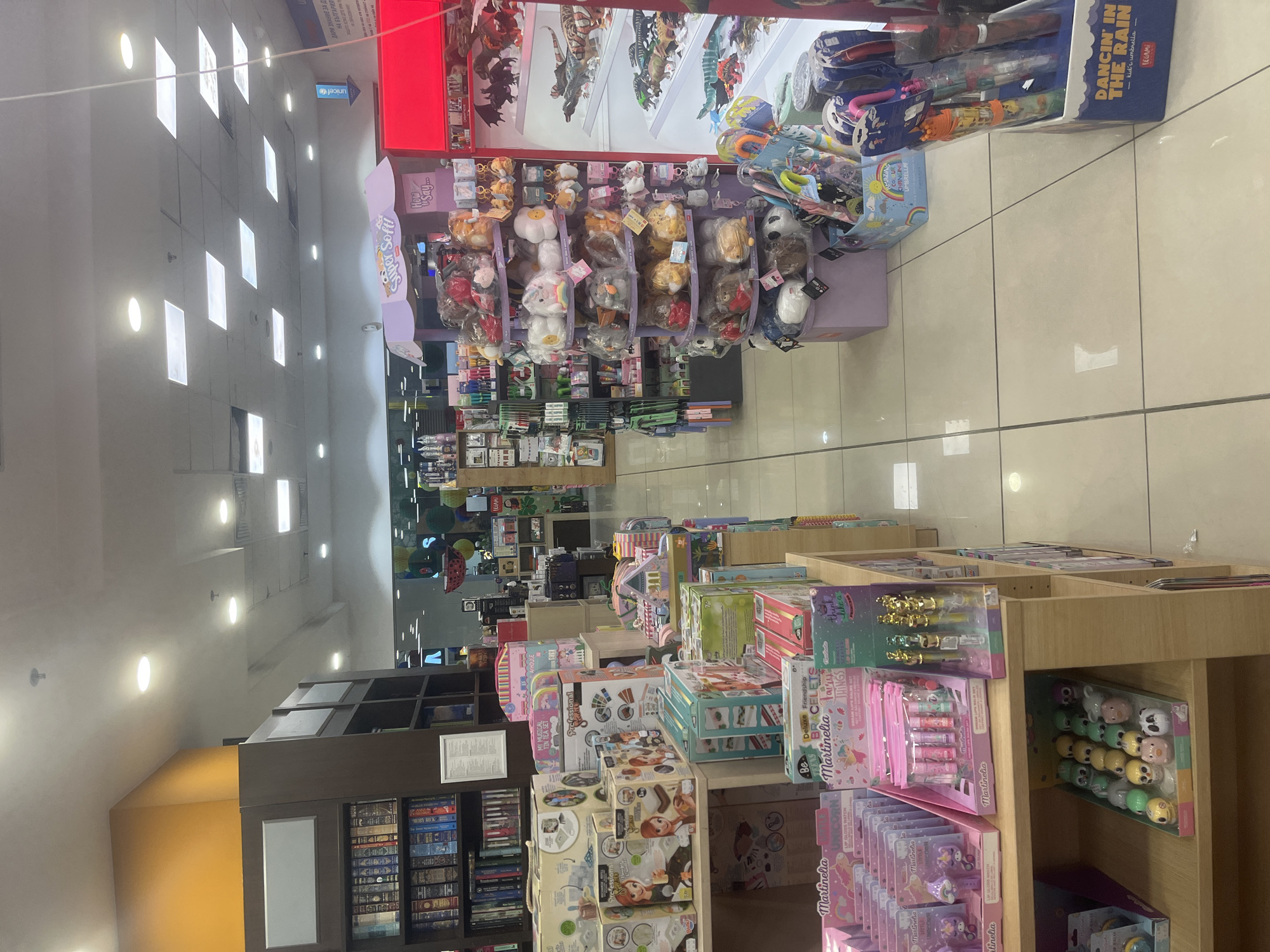 The image shows the interior of a store, likely a bookstore or gift shop, in Varna, Bulgaria, based on the "ДАРИ ЗНАНИЕ ЗА Д БЪЛГАРИЯ" sign, indicating a charity organization. The store is well-lit, with rows of shelves displaying various merchandise, including stuffed animals, toys, books, and novelty items. A prominent "Super Soft" sign can be seen in the distance, near a display of plush toys. Shelves closer to the camera feature items such as makeup sets, tea sets, and bracelet-making kits. The presence of the "unicef" sign suggests a partnership with or support for the organization. The scene appears to be organized and well-stocked.