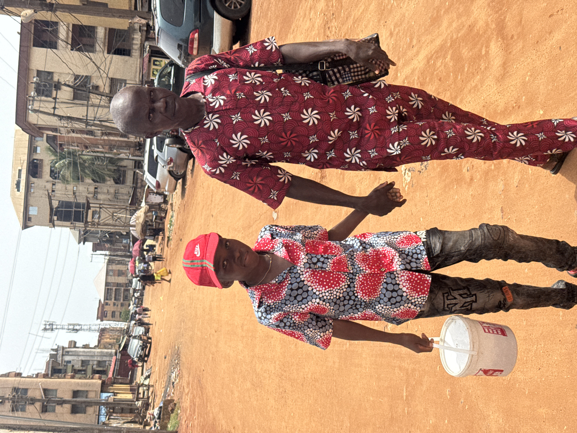In an outdoor scene in Onitsha, Nigeria, an older man in a long-sleeved red patterned outfit holds hands with a younger person wearing a red Nike cap, a short-sleeved shirt with a red and blue pattern, and jeans. The younger person carries a white bucket. They are standing on a dirt road in a busy market area. Buildings and power lines are visible in the background. Cars and pedestrians are on the sides of the road.
