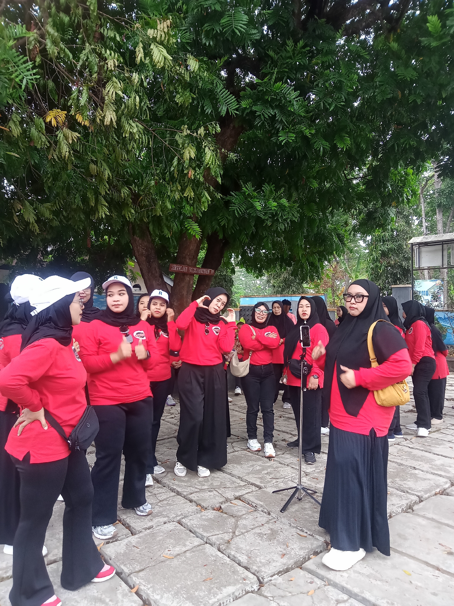 A group of women wearing red tops and black bottoms stand outside near a tree. The women are wearing hijabs. One woman in the foreground is wearing glasses and a yellow purse. A tripod is set up in front of the women. A sign on a tree behind the group reads "JALALUL NUROHIM". The ground is paved with large, flat stones. The sky is overcast. The location is identified as Kaligentong, Indonesia.