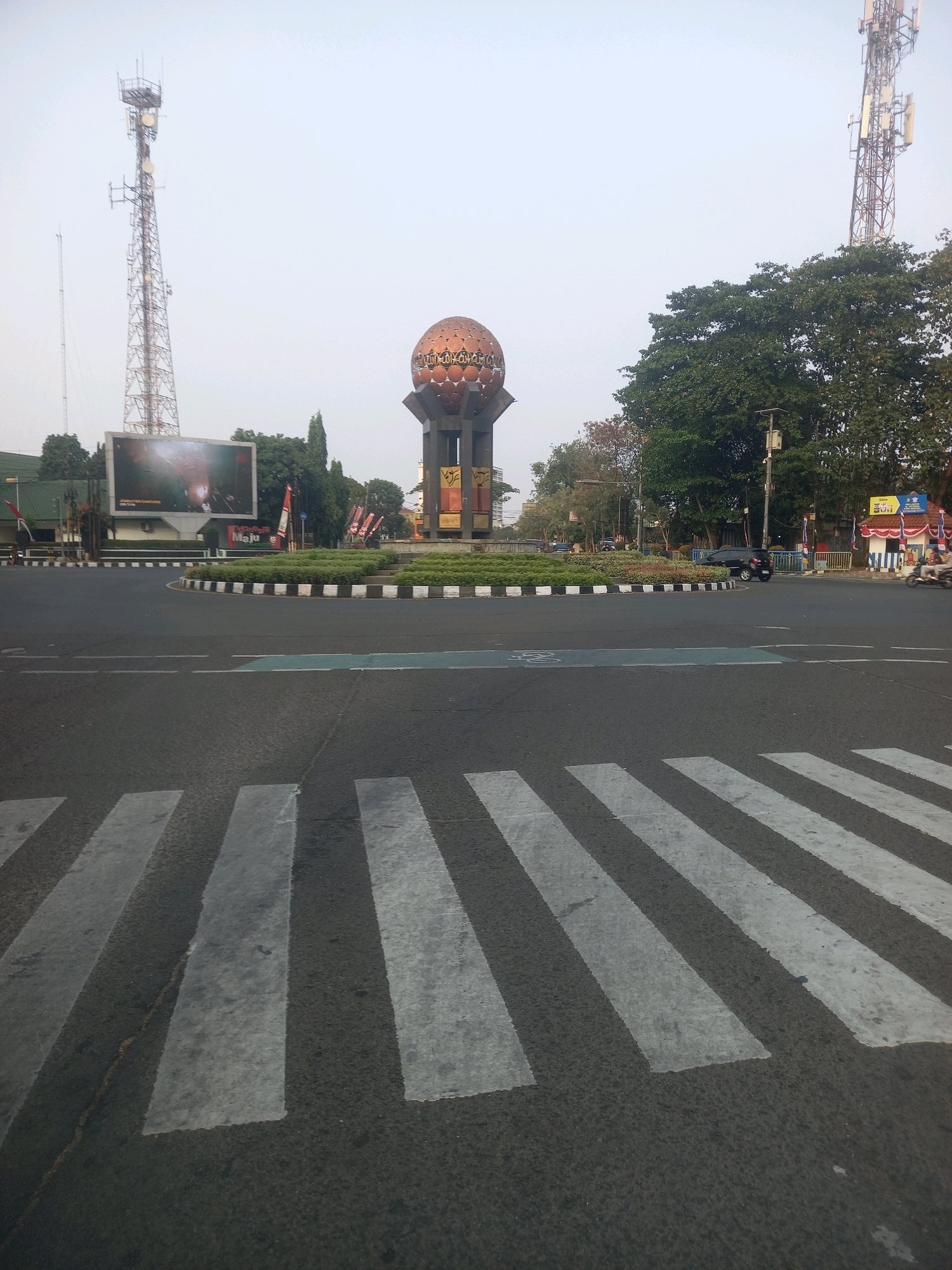 The image depicts a roundabout in Tangerang, Indonesia, featuring a large, ornate globe-like monument at its center, topped with what appears to be a golden, patterned sphere. Two prominent telecommunication towers flank the monument, one on the left and another on the right, extending high into the hazy sky. Below the left tower, a large billboard displays a vibrant image. The roundabout itself is lined with white and black curbs and a circular garden bed filled with greenery. A zebra crossing is visible in the foreground, painted with thick white stripes on the asphalt road. The road shows signs of traffic, with a black car navigating the roundabout and a scooter carrying two people to the right of the image. Trees with dense foliage surround the monument and border the road, adding to the urban landscape. The overall atmosphere suggests a bright, possibly early morning or late afternoon, due to the soft lighting and clear sky.