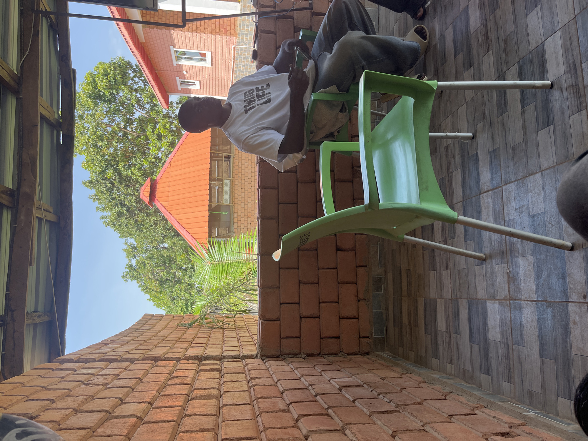 The image depicts an outdoor seating area with a man sitting in a green plastic chair. He is wearing a white t-shirt with the words "THUG LIFE" printed on it in black, and blue jeans. The man is looking to his right and appears to be holding something in his hands. In the background, there are brick walls, a partially visible orange-tiled roof, and lush green trees, suggesting a warm climate. The flooring is tiled with a wood-like pattern. The architecture and vegetation are consistent with a setting in Jos, Nigeria.