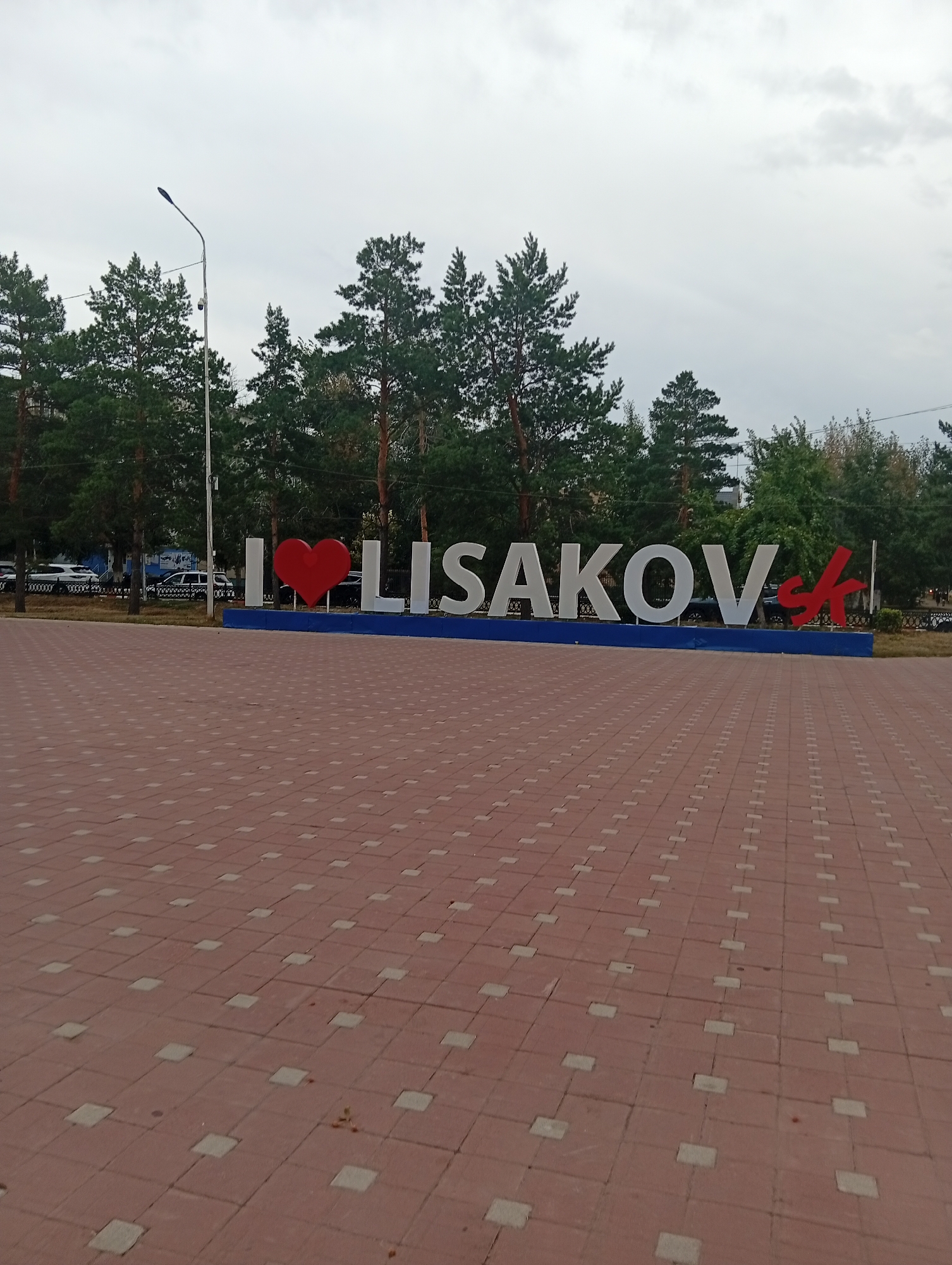 The image shows a large, three-dimensional sign that reads "I ❤️ LISAKOV". The sign is installed on a blue base and is situated in an outdoor urban setting. The foreground is dominated by a red brick-patterned pavement. Behind the sign, a row of tall green trees, likely pine trees, forms a backdrop. In the far background, there are buildings and cars. A street lamp with a camera attached is visible to the left of the sign. The sky is overcast. The sign is a landmark indicating the city of Lisakovsk, Kazakhstan.