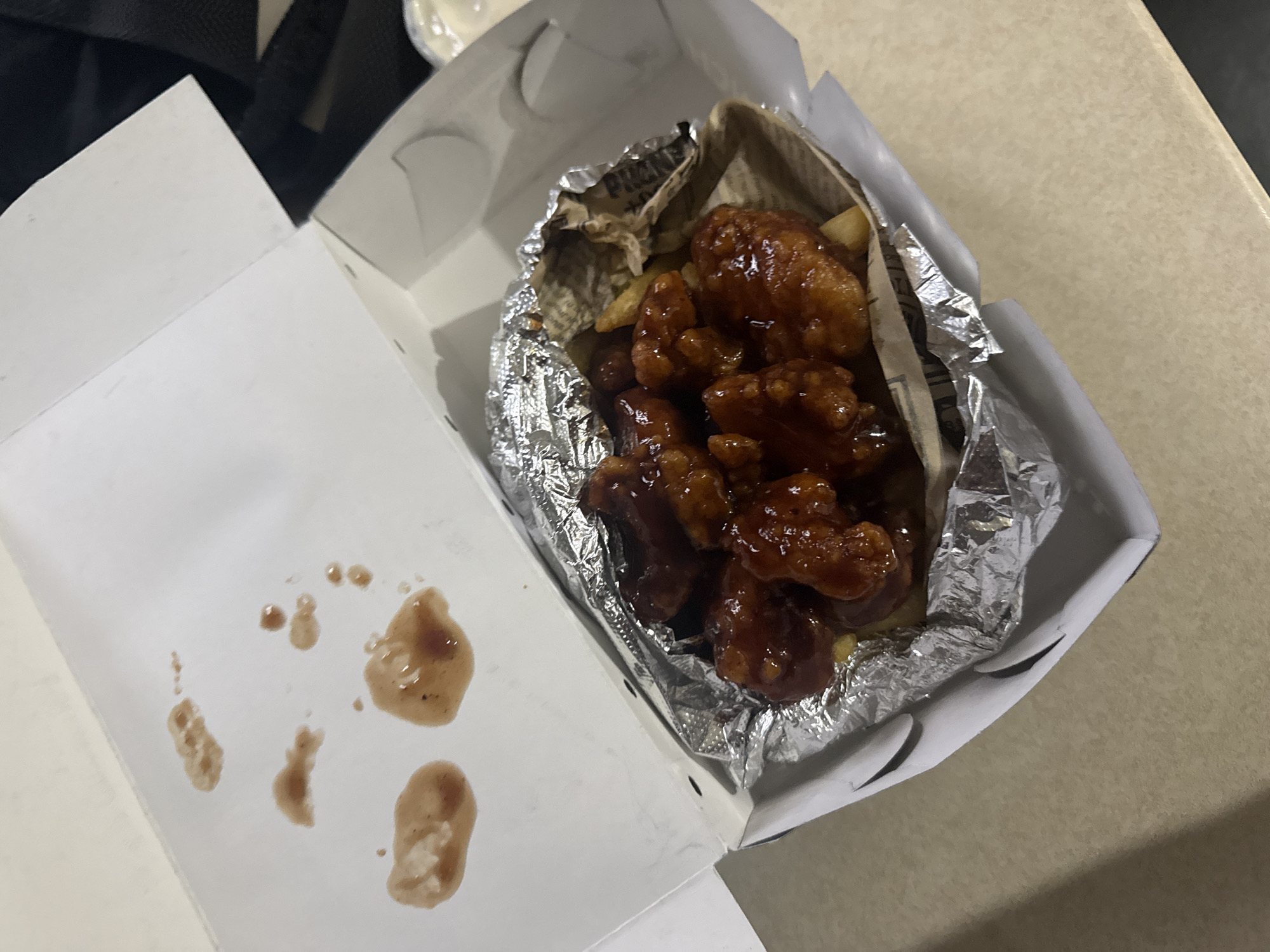 A high-angle shot depicts an open white cardboard takeout container, revealing several glazed, dark brown chicken wings nestled in aluminum foil. Some wings appear to have been partially consumed, and there are sauce splatters on the open lid of the container. A few french fries are also visible, mixed in with the wings. The photograph was taken indoors, likely on a table or countertop in an establishment in San Andrés Cholula, Mexico.