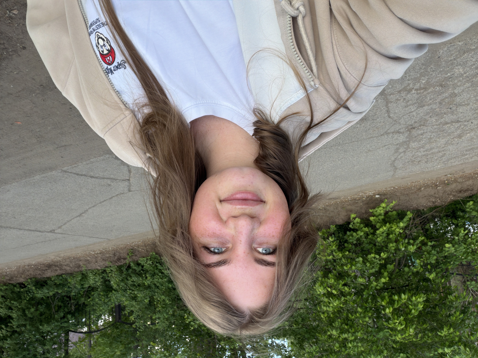 A woman is wearing a white t-shirt and a beige zipper hoodie with a Super Mario Brothers motif. The hoodie's design includes a cartoonish Super Mario wearing a red bandana. The location appears to be outdoors, with green foliage visible in the lower part of the frame. The upper portion shows a paved surface, likely a sidewalk or street. The picture is upside down, and no verifiable information ties the image to Chaikovsky, Russia.