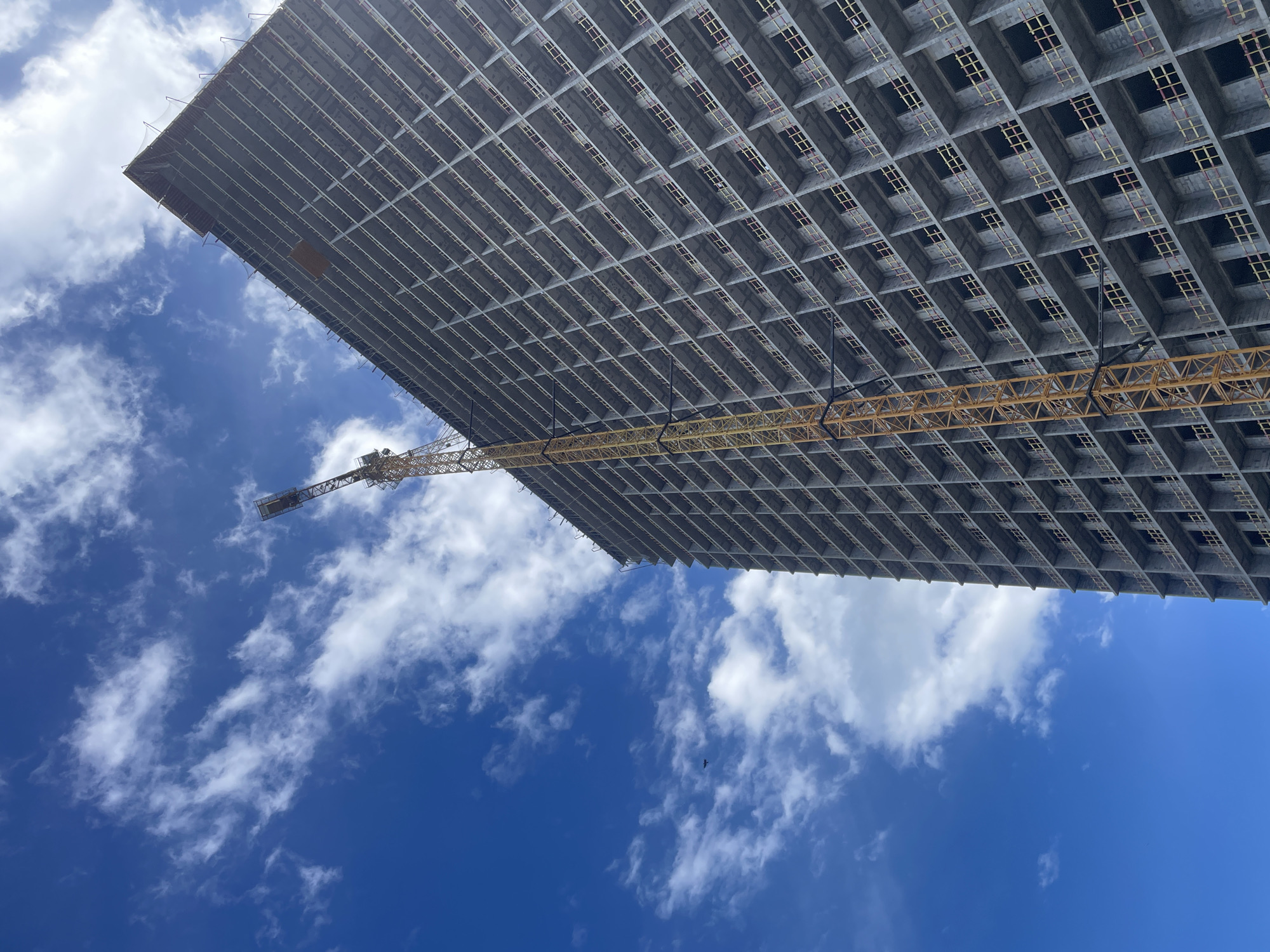 A low-angle shot depicts the top of a tall, unfinished building in Batumi, Georgia. The building's structure shows a grid of rectangular openings. A yellow construction crane extends from the top portion of the building. The sky is blue with scattered white clouds. No people are visible.