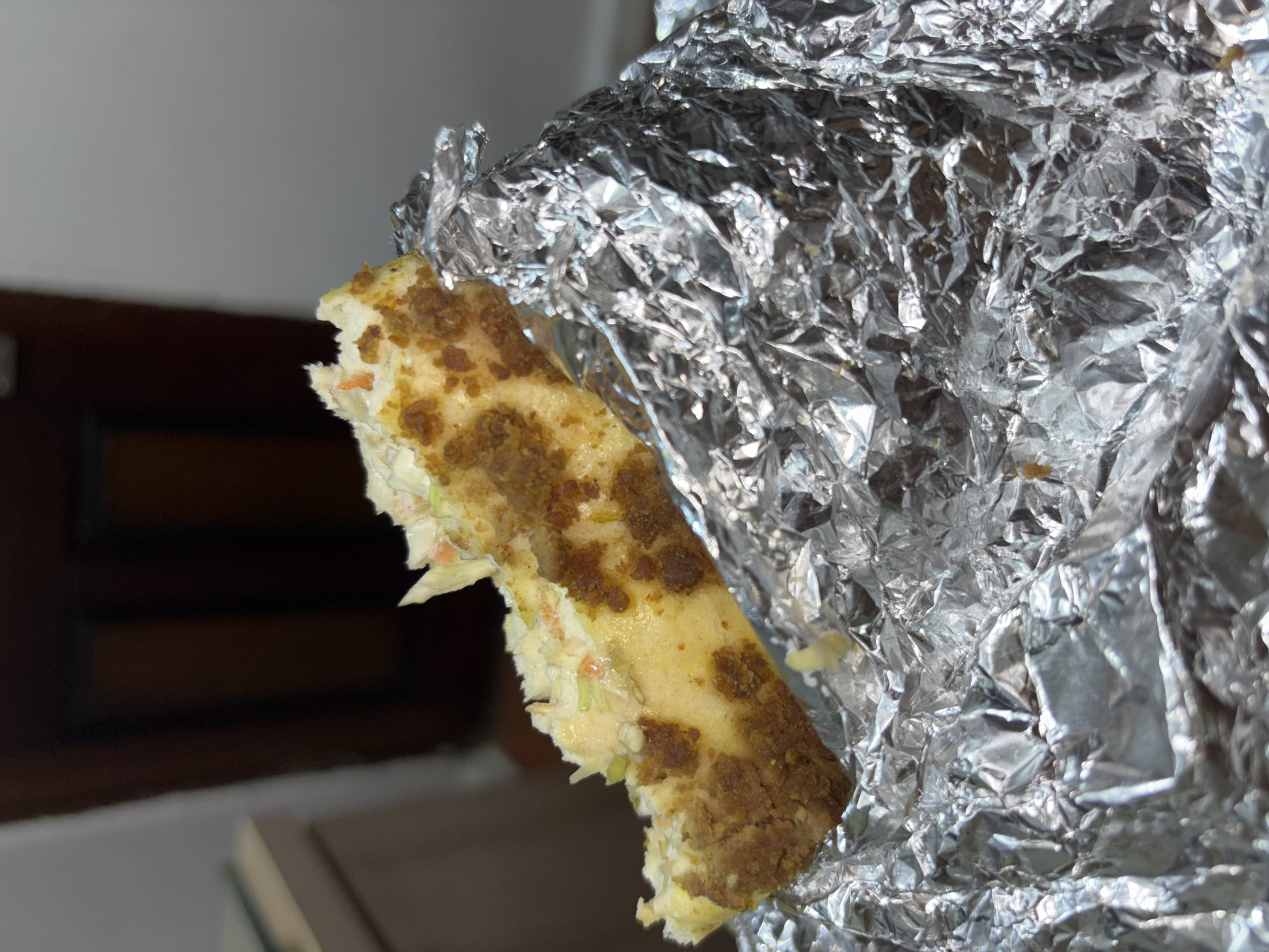 A partially eaten sandwich wrapped in aluminum foil is visible in the foreground. The sandwich appears to have a light-colored filling, possibly coleslaw, and is encased in bread with a textured, browned crust. The aluminum foil is crumpled and reflects light, creating a shiny, chaotic pattern. The background is blurred and indistinct, showing a dark-colored wooden structure and a light-colored wall. No people are visible. The location in Port Harcourt, Nigeria, is not identifiable from the image.