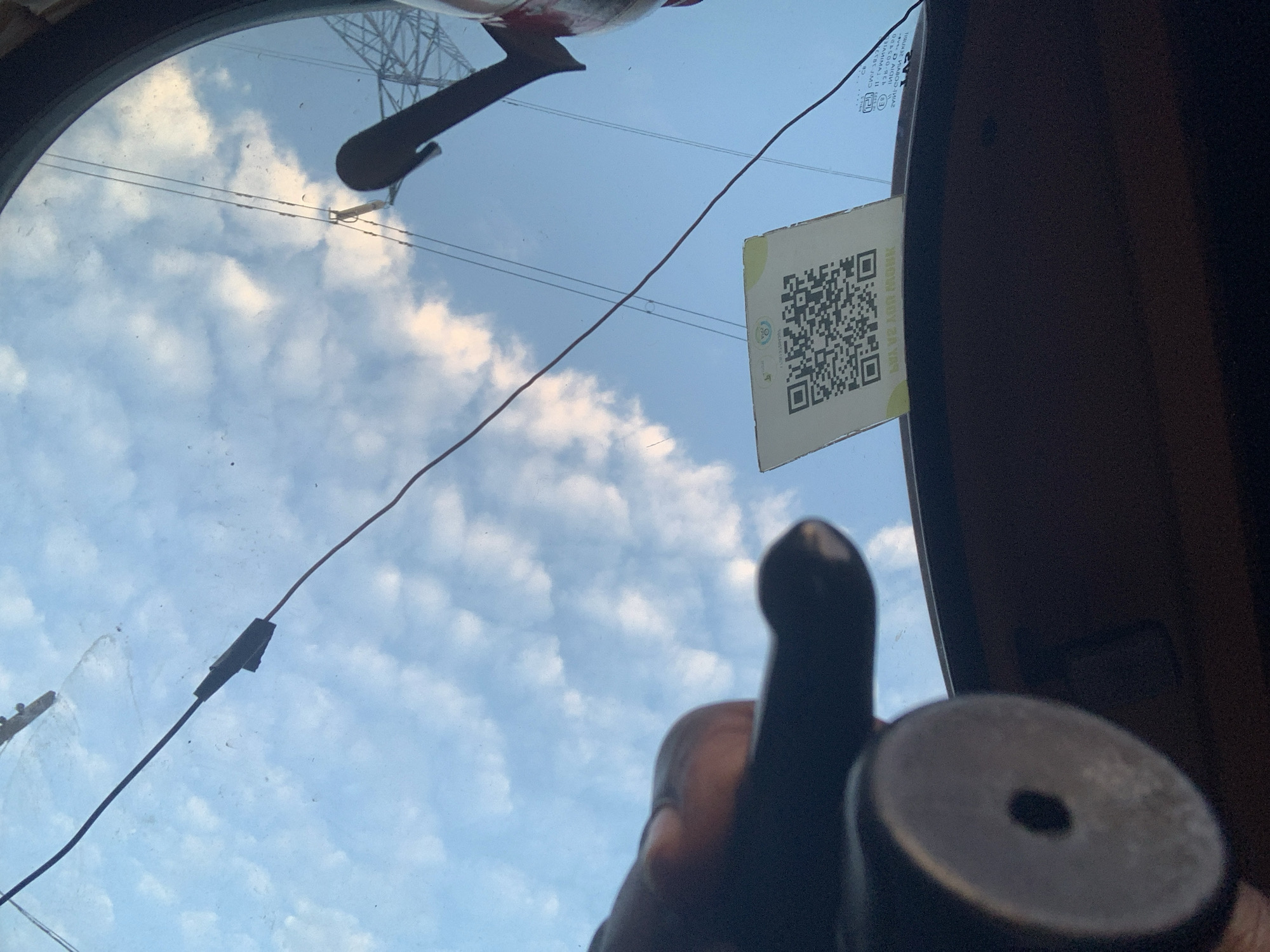 In Obosi, Nigeria, an upside-down view through a vehicle's windshield reveals a blue sky with scattered white clouds. A black object, possibly a part of the vehicle's interior or an accessory, is visible in the foreground, out of focus. A QR code sticker is affixed to the windshield, displaying a complex pattern of black and white squares. A thin, dark wire stretches across the windshield, attached to a small, dark grey connector. In the upper left of the frame, the silhouette of a power pylon is visible against the sky.