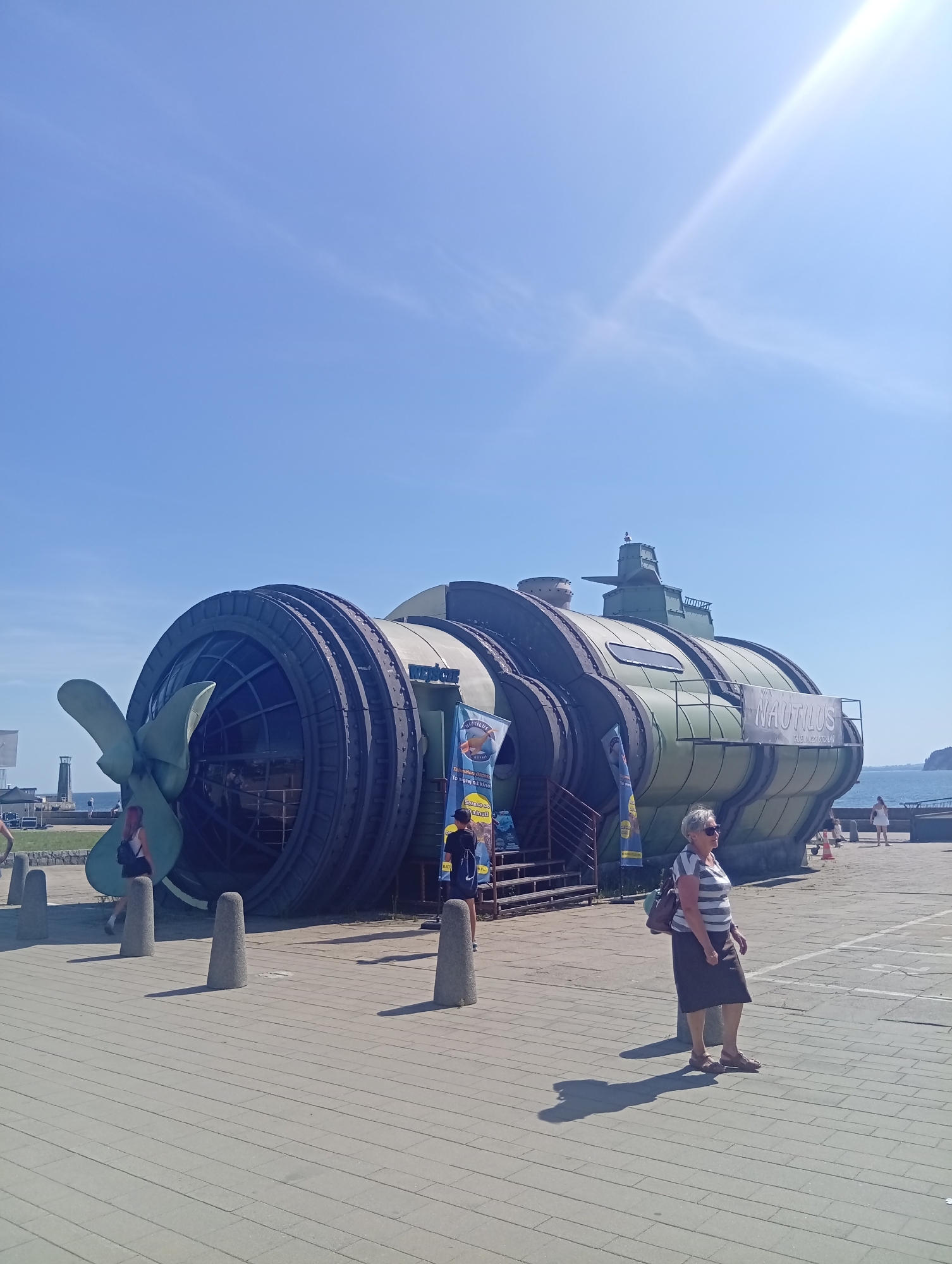 The image shows a large, green, submarine-like structure with a prominent propeller at the rear, resembling a tourist attraction or museum exhibit. The structure is labeled "NAUTILUS" on its side. It is situated on a paved area outdoors, with a clear blue sky overhead and the sea visible in the background. Several people are present, including a woman walking in the foreground wearing a striped shirt and dark skirt, and another woman in a white dress in the background. The submarine-like structure is likely located at a waterfront area in Gdynia, Poland, possibly near a pier or promenade, given the presence of the sea and the paved public space. The overall scene depicts a sunny day with a leisure-oriented atmosphere.