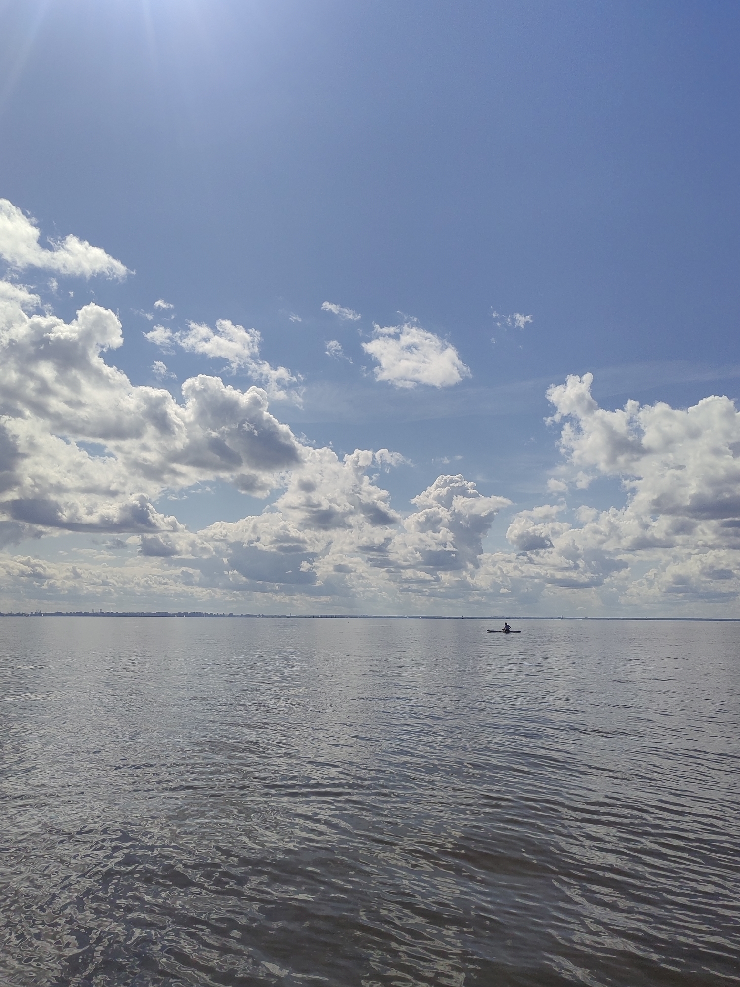 A lone figure paddles a kayak on the Neva River in Saint Petersburg, Russia, under a bright, partially cloudy blue sky. The river is calm with gentle ripples, reflecting the sky and clouds. In the distance, the Saint Petersburg skyline is visible as a faint silhouette along the horizon. The sun's rays create a bright glare in the upper left corner of the image.