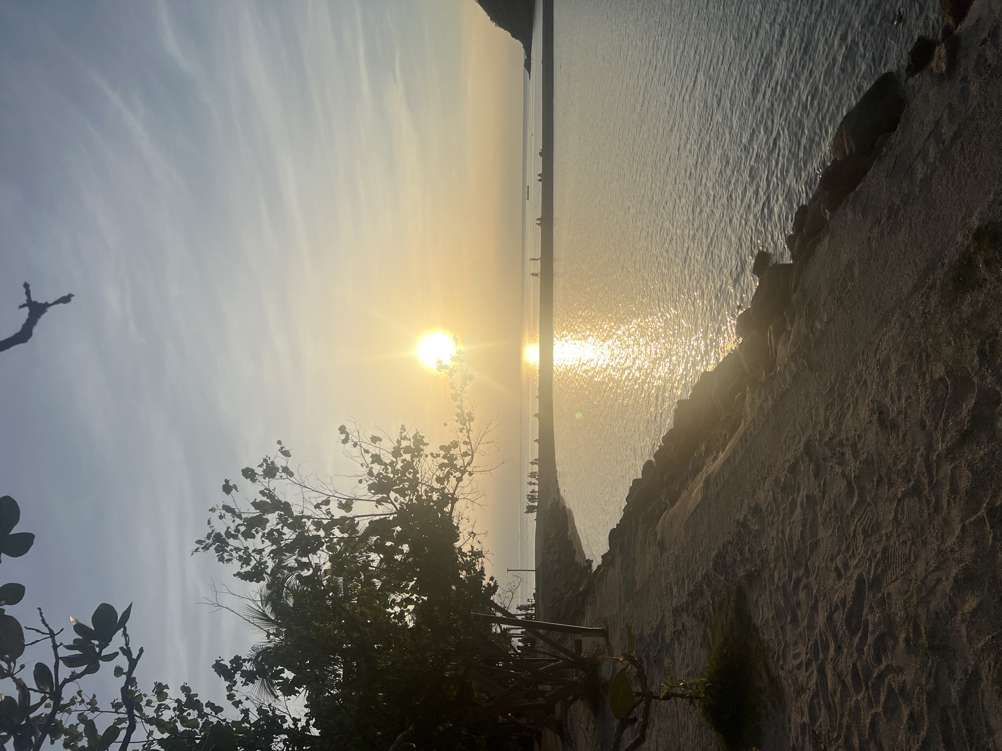 The image depicts a tranquil beach scene at sunset in Ko Pha-ngan District, Thailand. The sun, a bright orb with visible rays, is low in the sky, casting a warm golden glow across the water and the horizon. The sea is calm, with gentle ripples reflecting the sunlight, creating a shimmering path. A thin strip of land, possibly a sandbar or a narrow peninsula, stretches across the water, with several silhouetted figures standing or sitting on it. A few distant boats are visible on the horizon.
To the left, a sandy beach leads up to a line of rocks and then to the sea. Trees with dark foliage and visible branches are scattered along the edge of the beach, some of them silhouetted against the bright sky. There are also a few streetlights visible along the path bordering the beach. The sand has footprints, suggesting recent activity. The overall atmosphere is peaceful and serene, capturing the beauty of a tropical sunset.
