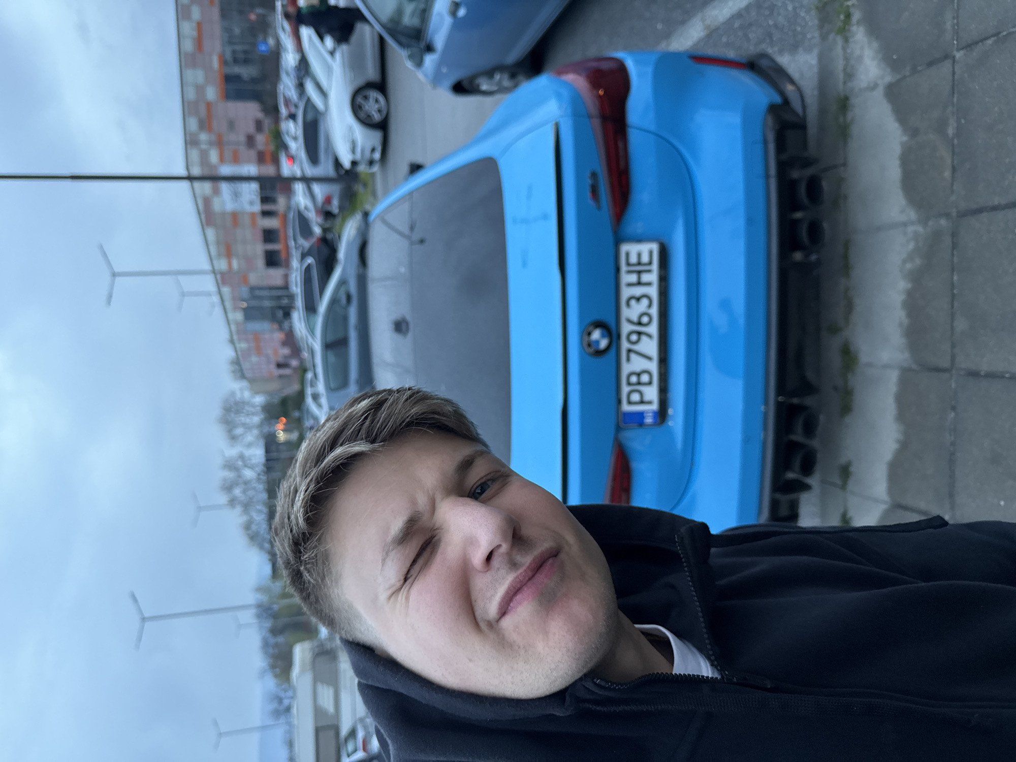 A man with short blonde hair is taking a selfie in front of a light blue BMW car with Bulgarian license plate "PB 7963 HE." He is wearing a black hoodie and is winking. The car is parked in a parking lot. Other cars are visible in the background. There is a building in the background with a pattern of orange and grey squares. There are lamp posts visible in the background. Based on the license plate, the location may be in Plovdiv, Bulgaria.
