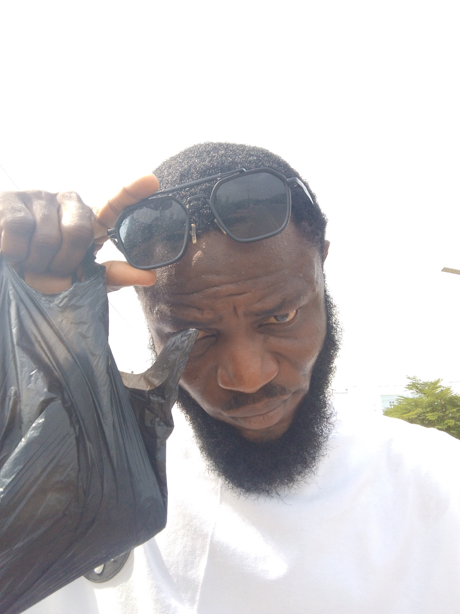 A person is holding a black plastic bag, touching their sunglasses with one hand, and squinting their eye. The subject has dark skin, a thick beard, and is wearing a white shirt. The location is possibly in Lokoja, Nigeria, but this is not definitively discernable from the image alone. Outside, there is a bright sky and a tree with green leaves visible in the background. There is a light pole or tall structure also visible.