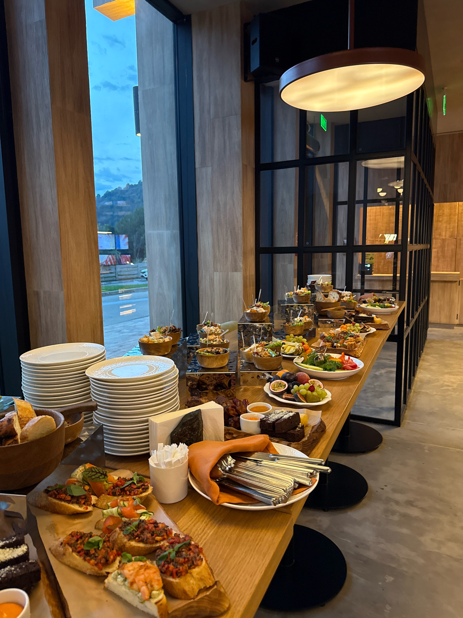 The image depicts a buffet table laden with various food items in a modern establishment, possibly a restaurant or hotel, located in Estosadok, Russia. The buffet spans a long wooden table, showcasing a diverse spread.
In the foreground, on the left, are stacks of white plates, some with decorative rims, and a wooden bowl filled with crusty bread. Adjacent to the plates, a cutting board displays several bruschetta topped with a dark, finely chopped mixture and garnished with herbs. One bruschetta is topped with a single shrimp and a creamy spread. To the right of the bruschetta is a white plate holding two square pieces of dark chocolate cake, a small yellow wedge, and two small white bowls containing orange sauce.
Further back on the table, an array of small bowls and platters hold various appetizers. These include small salads with visible vegetables and proteins, fruit platters with grapes, berries, passion fruit, and figs, and what appear to be mini quiches or tartlets. Several individual servings of appetizers, each with a toothpick or skewer, are arranged in wooden bowls.
A prominent feature of the scene is a large, round, pendant light fixture with a warm glow hanging from the ceiling. The background reveals a large window overlooking a landscape with hills and a cloudy sky, suggesting an outdoor view. The interior features wooden paneling on the walls and black-framed partitions, creating a contemporary aesthetic. The floor is a polished concrete. The overall impression is one of a well-prepared and elegantly presented buffet.