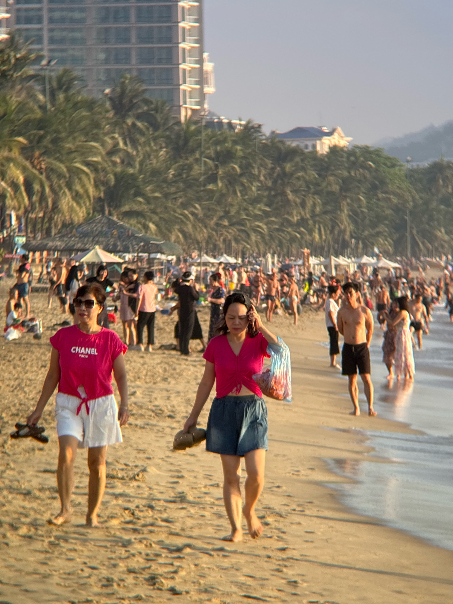 The image captures a bustling beach scene in Nha Trang, Vietnam, likely during daytime with the sun casting long shadows. In the foreground, two women walk on the sandy shore. The woman on the left, wearing a pink "CHANEL PARIS" t-shirt and white shorts, is holding sandals in her left hand. The woman on the right is wearing a pink top and denim shorts, carrying a plastic bag with items inside in her left hand. In the middle ground, numerous people are visible, some walking along the water's edge, others standing or interacting on the beach. Further in the background, palm trees line the shore, with resort-style buildings and a taller structure in the distance. The ocean waves are gently lapping at the beach, and beach umbrellas and shelters are scattered across the sand. The overall atmosphere is one of leisure and activity, indicative of a popular tourist destination.