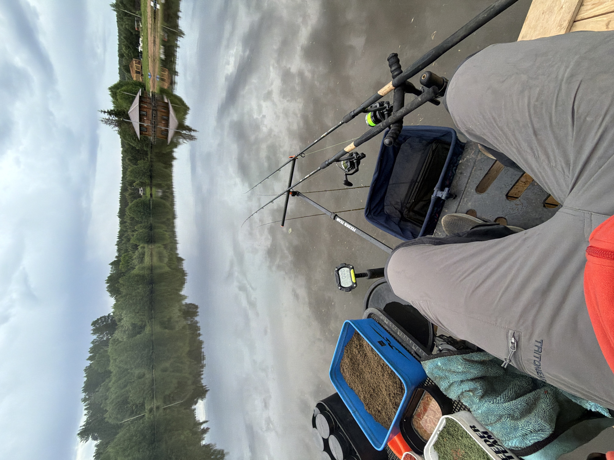 A person is fishing on a body of water in Prikamskii, Russia. Two fishing rods are set up. A blue tray with bait is visible, along with a blue keepnet. The shoreline with trees and a building are reflected in the water. The person is wearing gray pants.