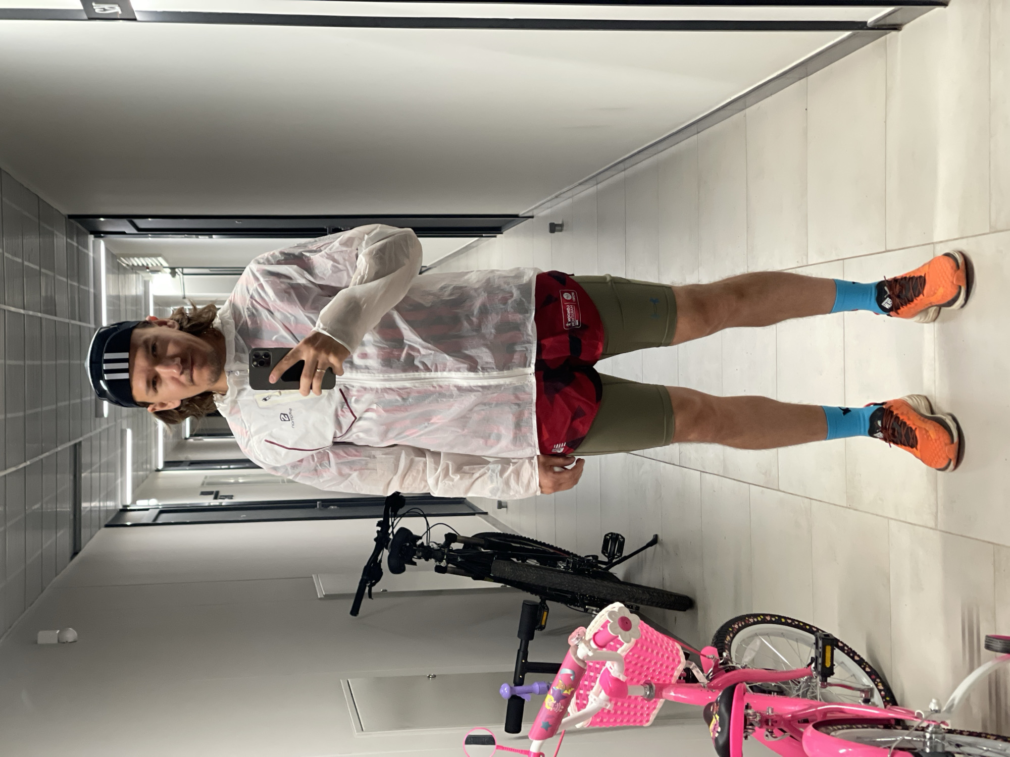 A person is standing in a tiled corridor with two bicycles to the left. The person is fair-skinned with brown hair and is wearing a black and white Adidas cap, a white and red windbreaker, olive green athletic shorts, red checkered underwear, blue socks, and orange sneakers. The person is holding a cell phone in their left hand. To the left of the person are two bicycles, one black and one pink. The location is Izhevsk, Russia.