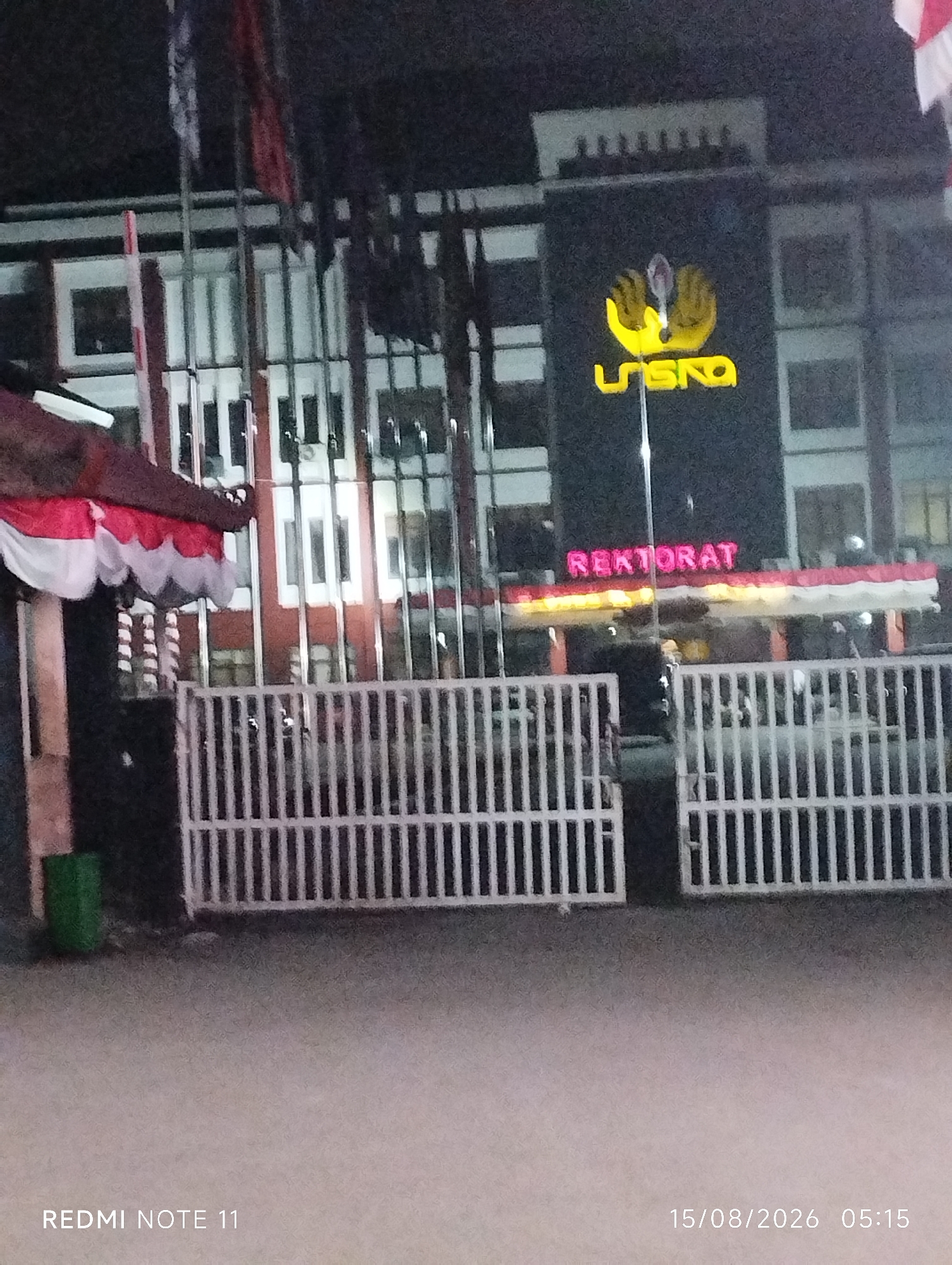 The image depicts the exterior of a building at night, likely a government or institutional building given the "REKTORAT" signage, which translates to "Rectorate" or "President's Office" in Indonesian. The building is dark, with illuminated signage. The prominent yellow logo of "UNSIKA" is visible, suggesting this is a building belonging to Universitas Singaperbangsa Karawang. The "REKTORAT" text is in red neon lights, positioned above a decorative banner with red and white stripes. A white metal fence runs across the foreground, partially obscuring the building's entrance. To the left of the fence, a green trash can is visible. The ground in front of the fence appears to be a paved surface, possibly a sidewalk or road. The overall lighting suggests it is nighttime, and the weather appears clear. The bottom right corner of the image displays a timestamp: "15/08/2026 05:15" and "REDMI NOTE 11". The location is identified as Karawang, Indonesia. There are no people or animals visible in the image.