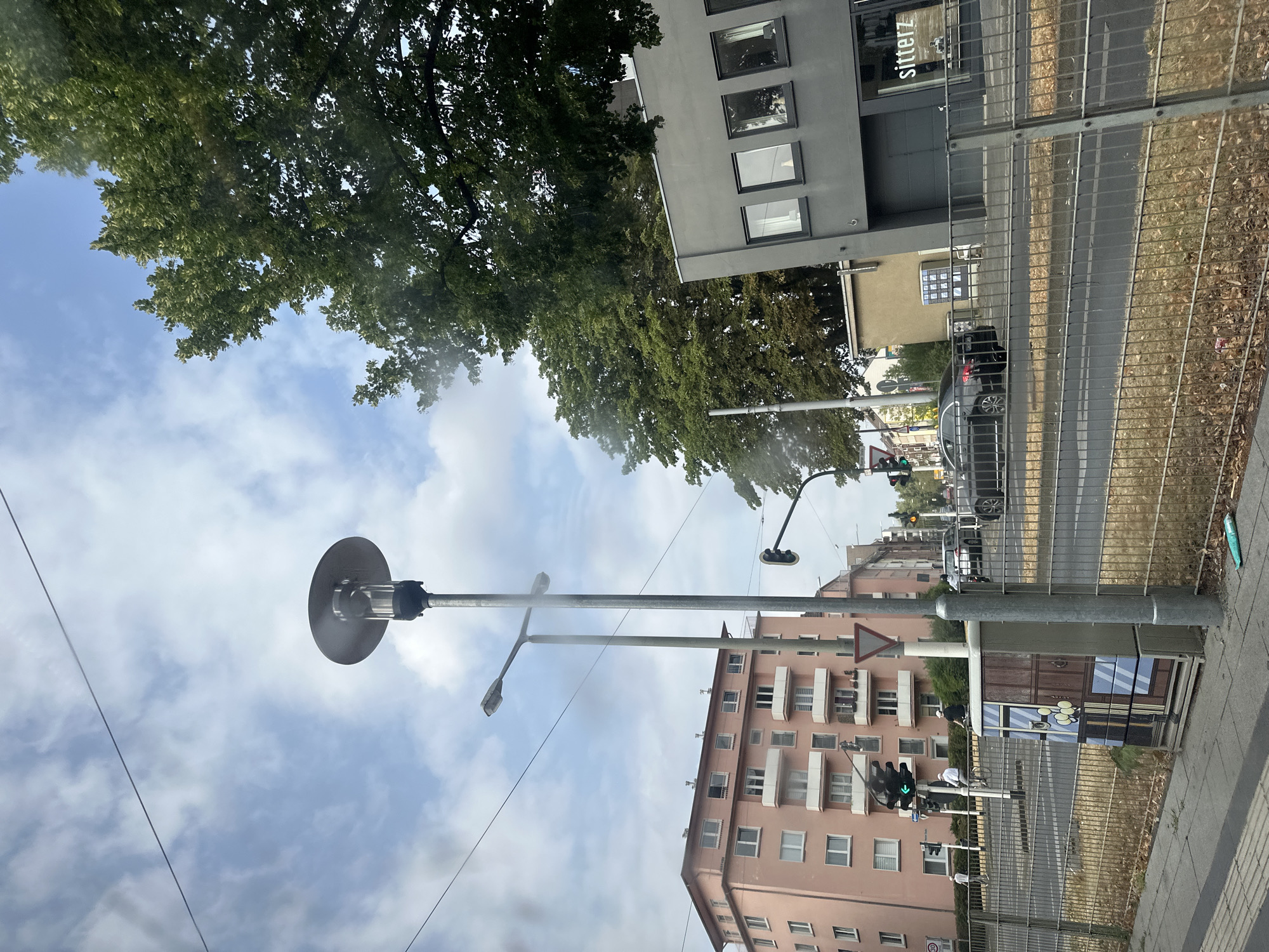 A street scene in Krefeld, Germany, features a pink apartment building with multiple balconies and windows. A modern grey building with large windows is on the right. In the foreground, a metal fence runs along the sidewalk. A street lamp stands tall, and traffic lights are visible. Several cars are on the road, and a few people are walking or cycling. Trees with lush green foliage are on the right side of the image. The sky is cloudy.