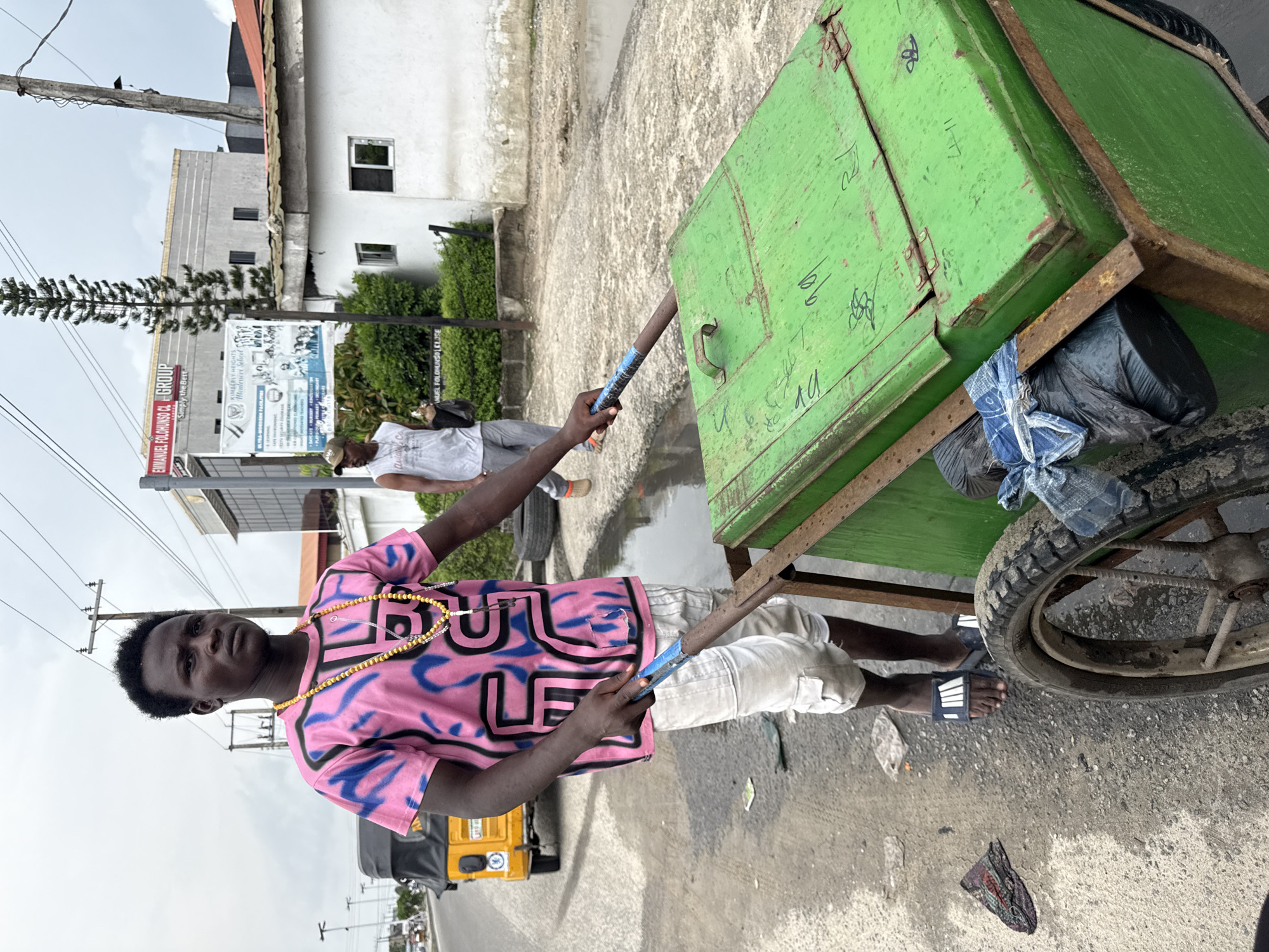 A young man of African descent is pictured from the waist down, pushing a green wheeled cart on a wet, paved street in Ado, Nigeria. The man is wearing a pink t-shirt with blue graffiti-style lettering, white cargo shorts, and dark sandals with white stripes. He has yellow beads around his neck and is holding onto the cart's handle with his right hand. The cart has a green lid with some handwritten markings and appears to contain trash bags. In the background, a yellow tricycle is visible on the street, and buildings, including one with a red sign, line the road. The sky is overcast.