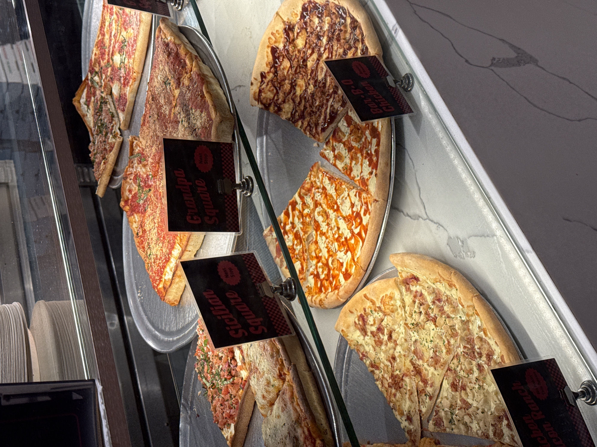 The image shows a display case filled with various types of pizza slices. There are at least four different pizza varieties visible: "Grandpa Square," "Sicilian Square," "Bar-B-Q Chicken," and "Chicken Bacon Ranch." Each pizza type has a small sign identifying it. The slices are arranged on metal trays inside the glass-fronted display case. The environment is likely an eatery or pizzeria in New York City, United States of America.