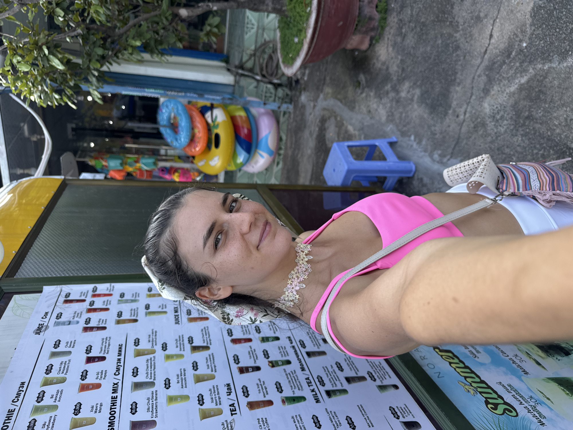 A woman with brown hair and a pink top stands in front of a menu board outside a shop in Mũi Né, Vietnam. The menu board displays various drinks, including smoothies, juices, tea, and milk tea, with prices listed in Vietnamese Dong and Russian. In the background, inflatable rings are stacked, and a potted plant is visible. The ground is wet, suggesting recent rain. The woman is holding a camera, taking a selfie, and wearing a decorative choker necklace.
