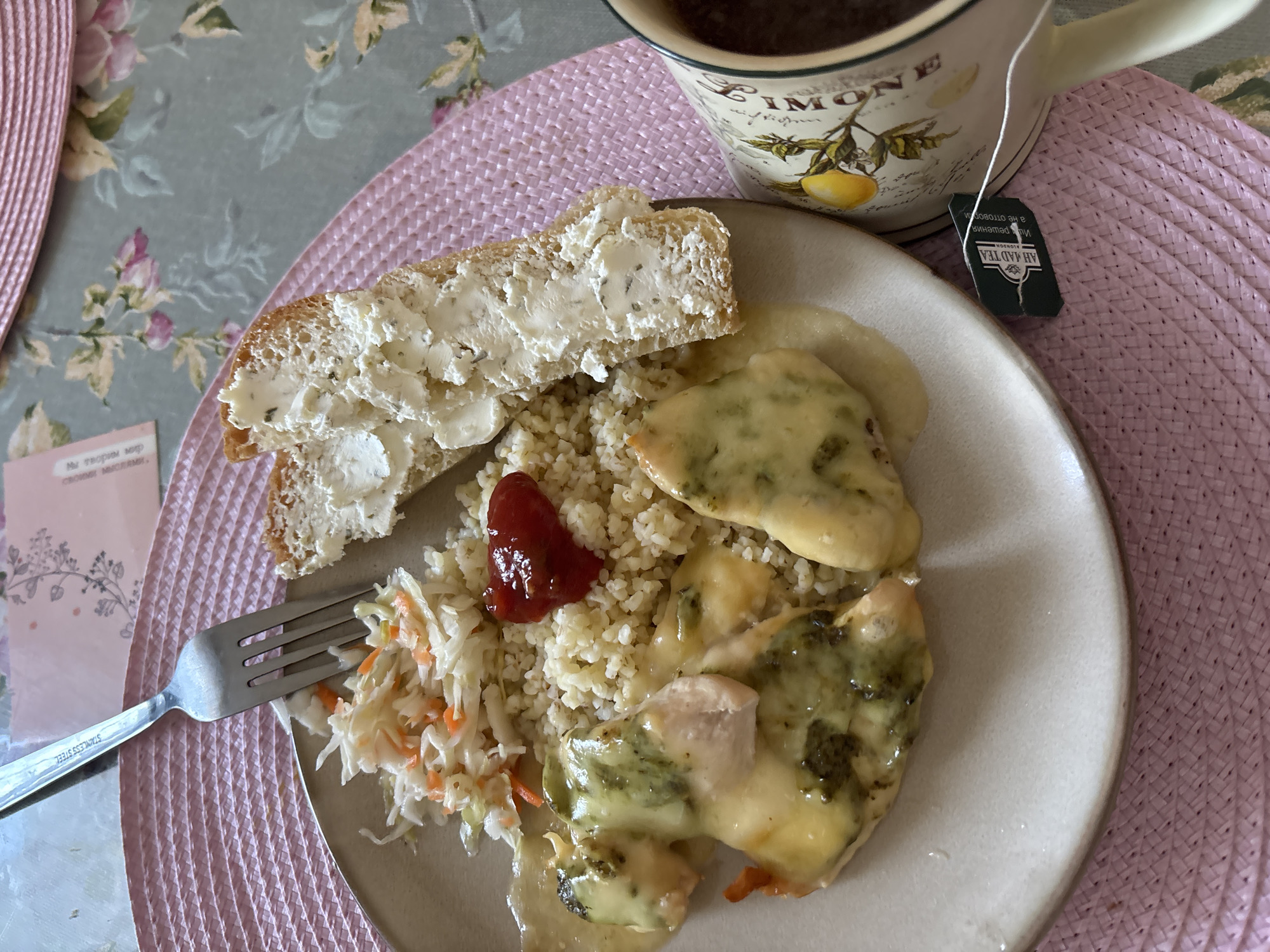 A close-up, overhead shot captures a meal on a plate, possibly for breakfast or lunch, set on a pink woven placemat. The meal consists of bulgur, coleslaw, and chicken coated in a creamy sauce and herbs, accompanied by a slice of bread with butter and a dollop of ketchup. A mug of dark liquid, likely tea or coffee, sits to the right, with a tea bag tag visible. The background features a floral tablecloth, suggesting an indoor setting. The image lacks any discernible indicators of its specific location in Naberezhnye Chelny, Russia.