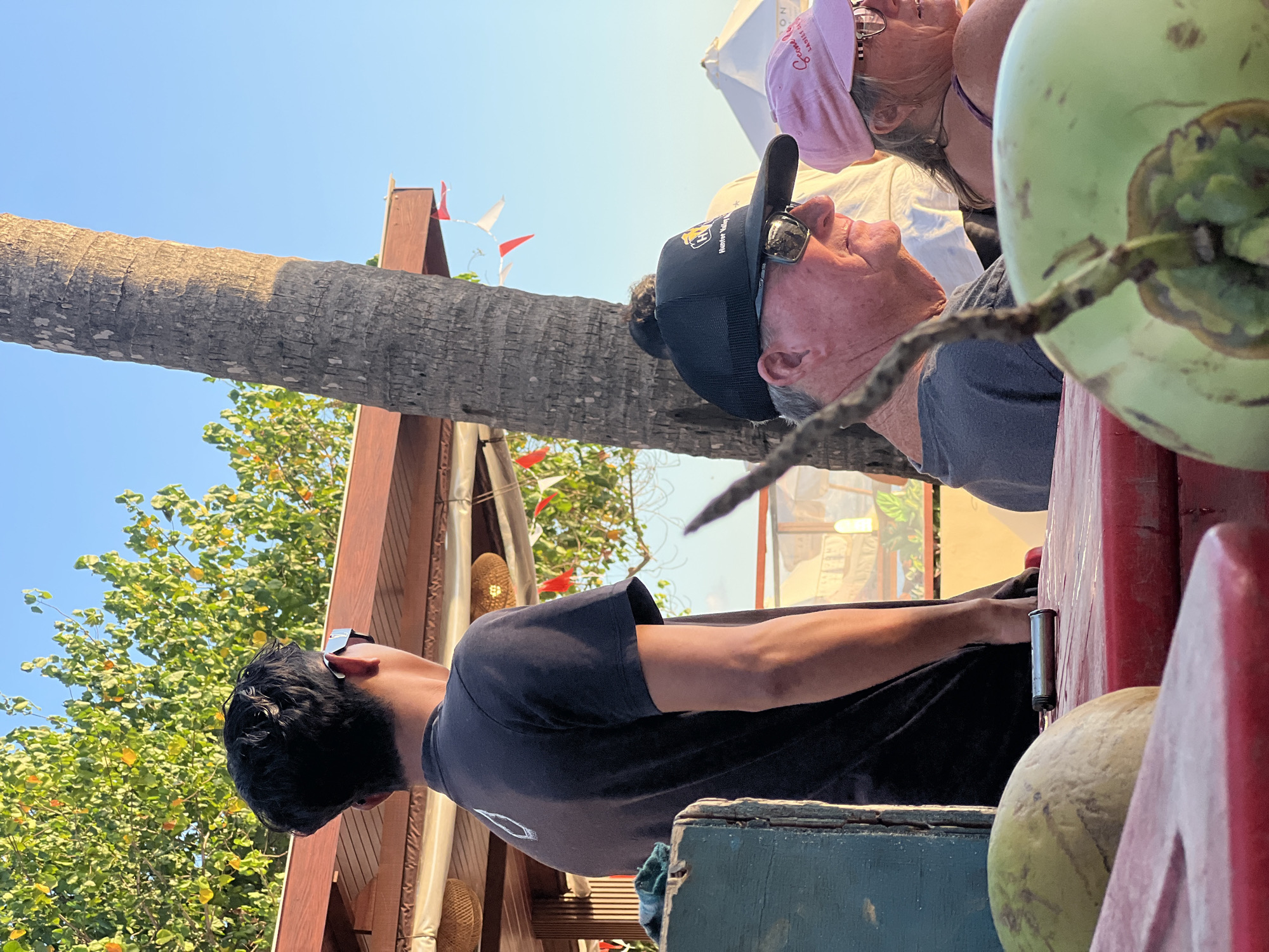 In Kuta, Indonesia, a young person in a black t-shirt stands facing away from the camera, looking towards a tree. In the foreground, a man wearing sunglasses and a black baseball cap with "Hunter Valley" visible on it, is seated and looking to his right. Beside him, a woman in a pink hat and glasses is also seated and looking to her right. Two green coconuts are in the foreground, one in focus and the other blurred. The background shows a palm tree, a thatched roof structure, and foliage under a clear blue sky.