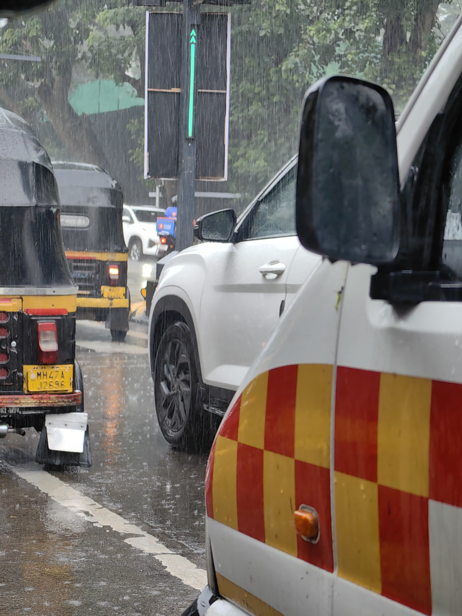 The image captures a street scene in Mumbai, India, during a heavy downpour. In the foreground, the side of a white vehicle with a red and yellow checkered pattern is visible, along with its orange indicator light. Partially visible in the left foreground is the rear of a black and yellow auto-rickshaw with a yellow license plate reading "MH 47 A J 2696". Further back, another auto-rickshaw is seen, and behind it, a white car. The road is wet and reflects the surrounding lights. A traffic light displaying a green arrow pointing upwards is also prominent in the scene, indicating that traffic can proceed in that direction. The overall environment is characterized by rain and wet surfaces, suggesting a monsoon season or heavy rainfall.