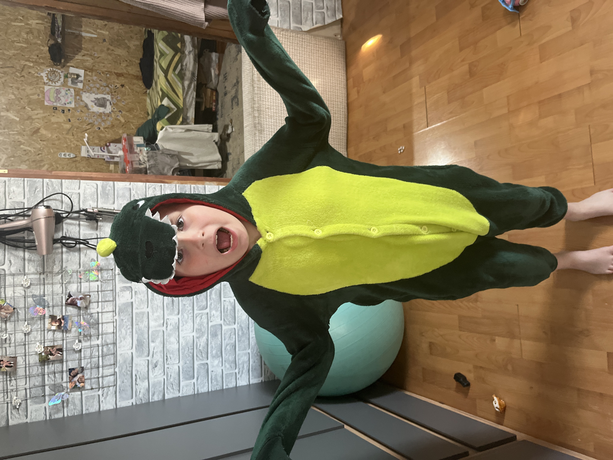 An indoor, vertical shot taken in Vesyoloye, Russia, captures a young child, likely a boy, in a playful moment. The child is wearing a full-body, dark green and bright yellow dinosaur kigurumi or pajamas, featuring buttons down the front and a hooded head with white teeth and a small yellow spike. His mouth is open in an exclamation or laugh, and his eyes are wide, conveying excitement and playfulness. His bare feet are visible at the bottom right.
The setting appears to be a child's room or play area. The background features a textured wall on the left, composed of a light-colored wooden panel adorned with various children's drawings and stickers. A hairdryer hangs near the top left of this panel. Below it, a grey brick-patterned wall holds a metal grid displaying small photographs and iridescent butterfly decorations. A dark-colored furniture piece, possibly a low bed frame, runs along the bottom of the image.
On the right side, the floor is made of light wooden planks. A large, light blue exercise ball is partially visible behind the child. Further back, a bed with a green, yellow, and white patterned comforter can be seen. Two small toy figures, one black and one orange and white, are scattered on the wooden floor. There is no specific time of day indicated, but the lighting suggests daytime. The only visible text, "DDD," appears on a small red object on the wooden wall panel. The overall scene suggests a vibrant and active home environment.