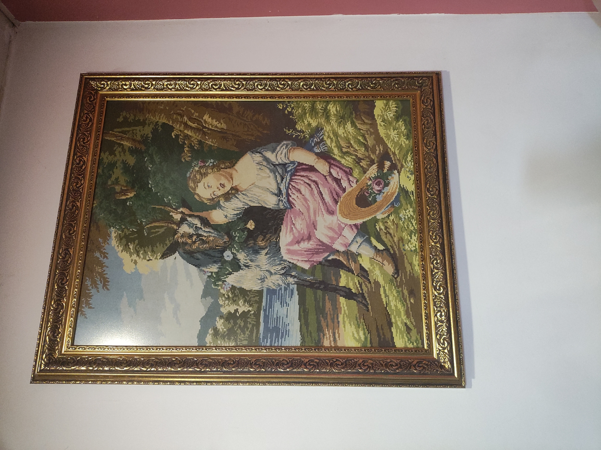 A framed tapestry hangs on a white wall with a pink trim in the city of Unknown, Unknown. The tapestry depicts a woman with fair skin and blonde hair, wearing a light blue and pink dress, lying on the back of a goat. She is holding a straw hat with flowers in it. The goat is gray and white. The background shows a landscape with trees, a waterfall, and a green field. The frame is gold with an ornate pattern.