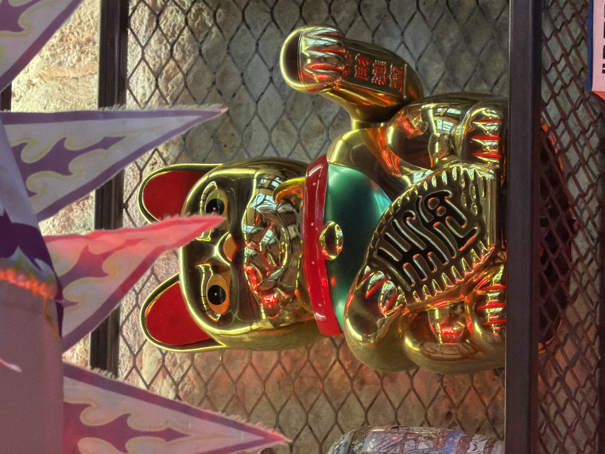 A brightly lit, close-up shot presents a golden Maneki Neko, or fortune cat, positioned behind a wire fence against a textured wall. The cat is partially obscured by triangular flags decorated with flame-like patterns in white and purple. The fence and wall, along with the reflections on the shiny golden cat, indicate an indoor setting likely within a shop. The photograph was taken in the city of Paris, France.