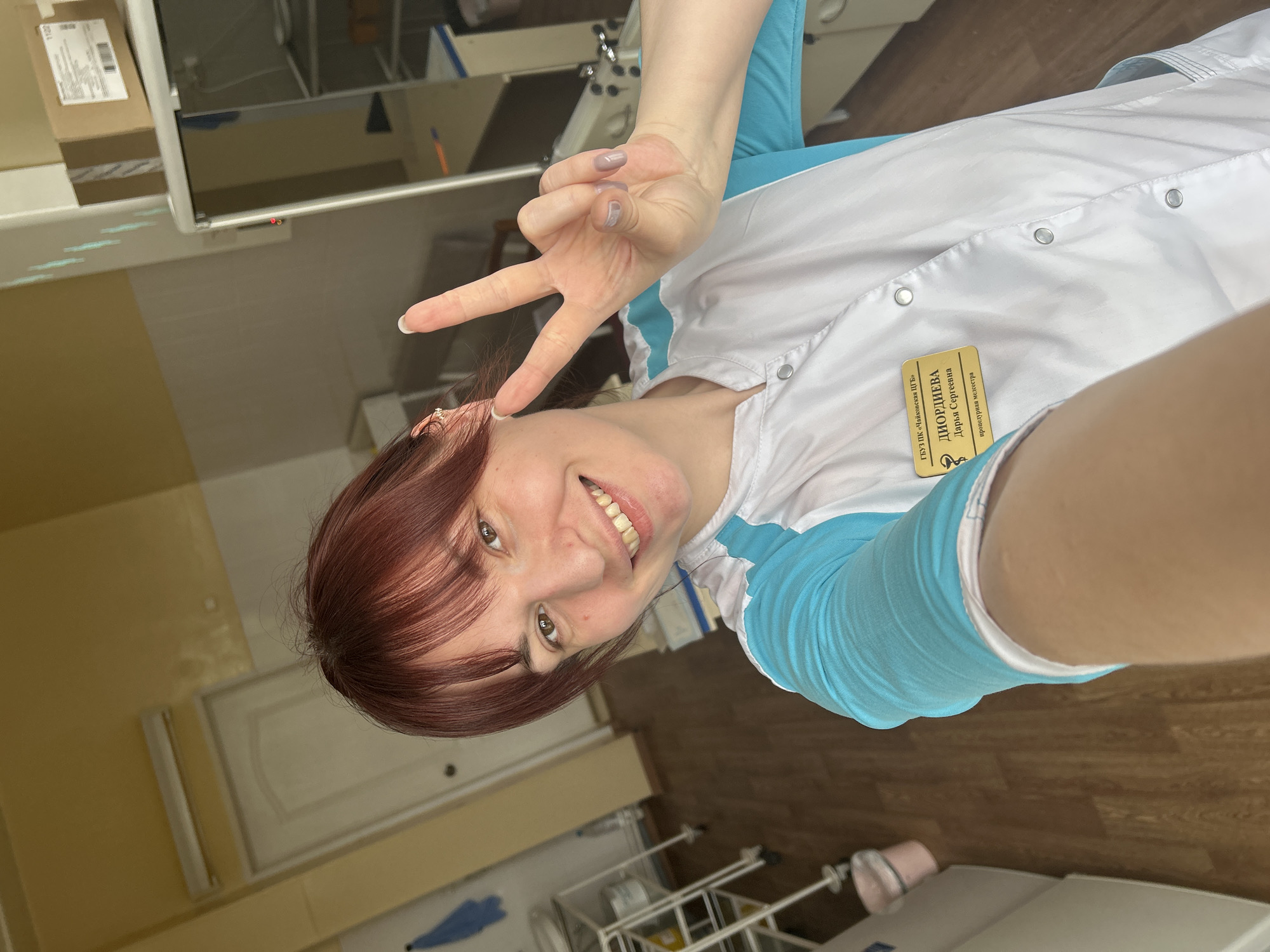 A woman with reddish-brown hair, wearing a white coat with light blue sleeves, is making a "peace" sign with her hand. She is smiling at the camera. The name tag on her coat reads "ДИОРДИЕВА Дарья Сергеевна" and "ГБУЗ ПК «Чайковская ЦГБ» процедурная медсестра". The background suggests an indoor environment, possibly a medical facility, with light yellow walls and medical equipment visible. Based on the name tag, this is likely in Фоки, Russia, as "ГБУЗ ПК «Чайковская ЦГБ»" indicates Chaykovsky Central City Hospital.