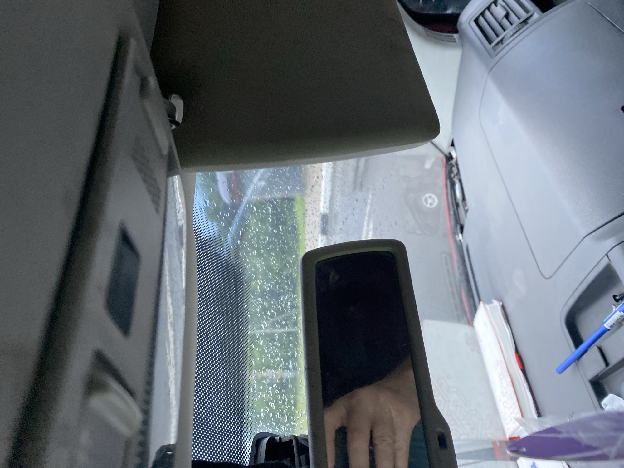 The image shows the interior of a vehicle, looking towards the windshield and dashboard. It is raining outside, with raindrops visible on the windshield and in the background. A rearview mirror is mounted above the dashboard, and a sun visor is visible on the right side of the frame. The dashboard itself is cluttered with various items, including a book, a pen, and a purple object. A vent for the car's climate control system is visible on the right side of the dashboard. The overall scene suggests a person is inside a parked vehicle on a rainy day. The location is identified as Unknown, Unknown.