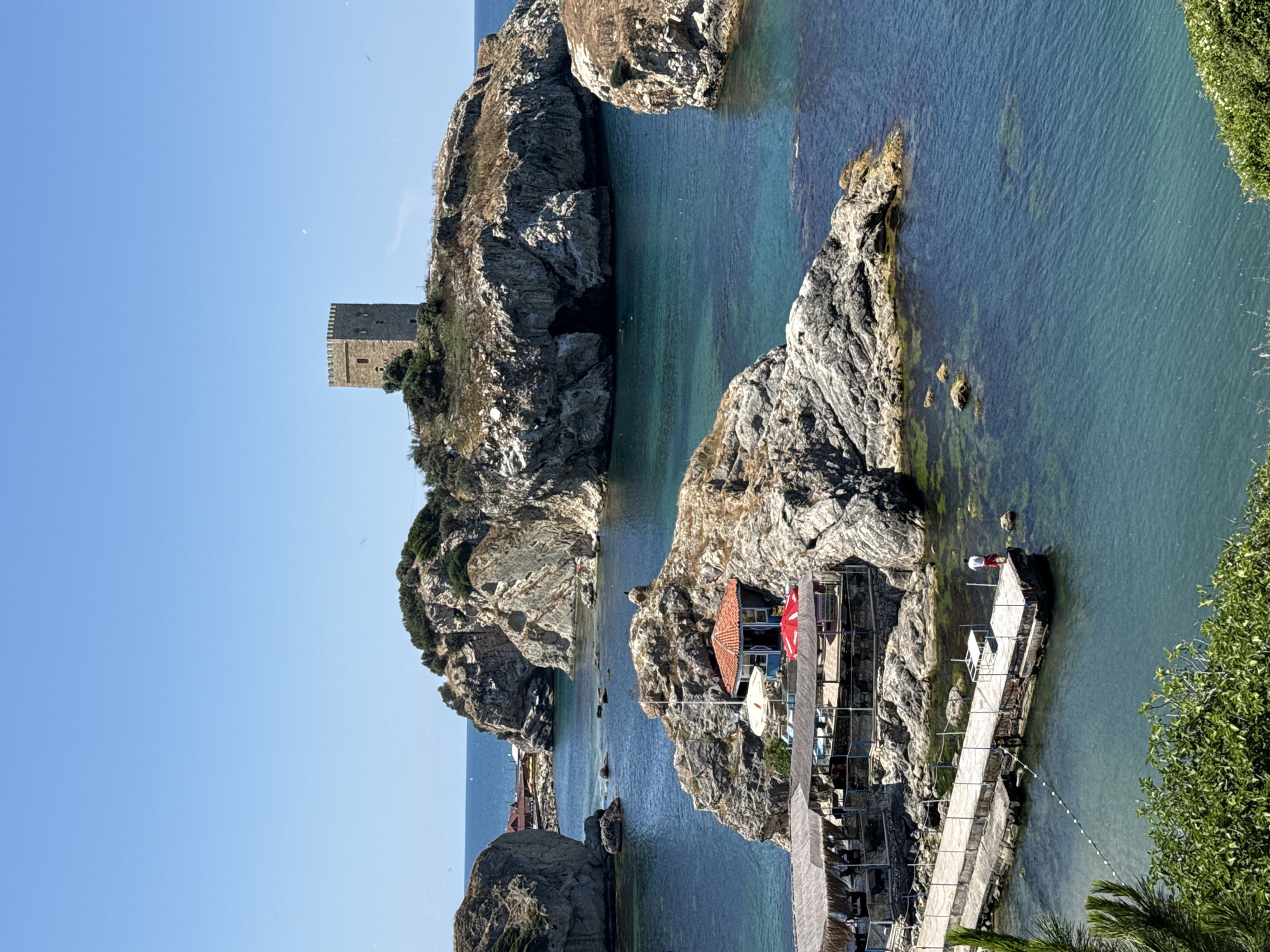 The image depicts a scenic coastal view in Şile, Turkey, featuring rugged rock formations jutting out of the clear turquoise sea. On the largest island, a stone tower with battlements stands prominently. A small, red-roofed structure with blue walls is nestled on a rocky outcrop closer to the foreground, adorned with two umbrellas, one white and one red. Adjacent to this, a wooden pier extends into the water, with a person in a red shirt and white shorts standing on it, facing away from the camera. Sheltered seating areas with thatched roofs are visible on a rocky platform near the pier. The water is calm and transparent, revealing the rocky seabed and some patches of green algae. Lush green vegetation frames the lower part of the image. The sky is a clear, bright blue.