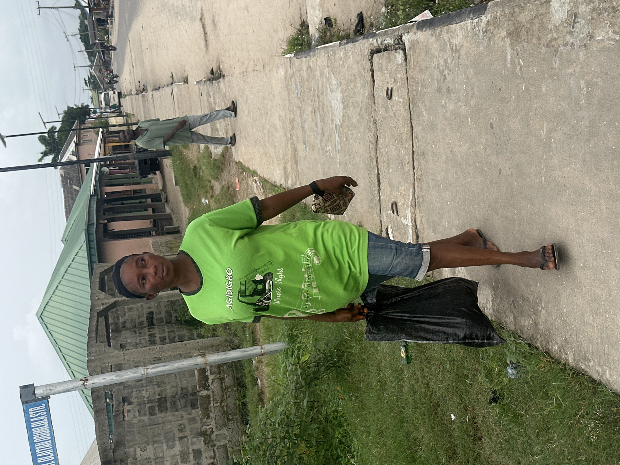 A woman in a bright green t-shirt and denim shorts walks on a concrete sidewalk in Badagry, Nigeria. She is carrying a black plastic bag in her left hand and a small patterned purse in her right hand. She wears black flip-flops on her feet. Behind her, a man in a green shirt and gray pants walks further down the sidewalk. The street has a paved road with several motorcycles and a white bus visible in the distance. The environment appears to be a commercial area with buildings lining the street, some with green roofs. A street sign reading "R. OLAITAN OGUNLOLA STR." is visible on the left side of the image. The ground is a mix of concrete, dirt, and grass. The sky is overcast.