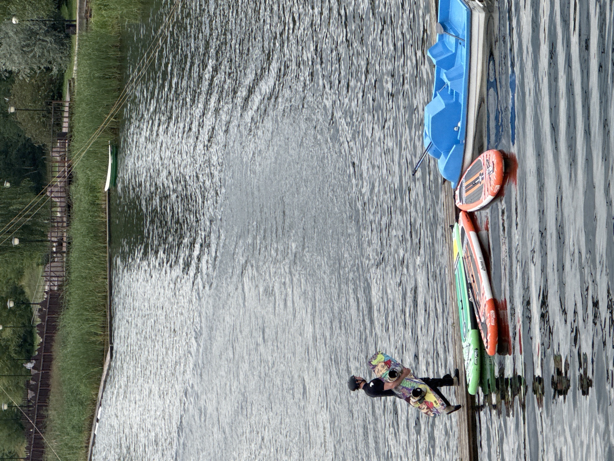 The image shows a man holding a wakeboard while standing on a wooden dock on a body of water. Beside the dock, there are two paddleboards, one green and one orange, and a blue pedal boat. In the background, there is a dense line of green reeds, a wooden fence, and trees. The sky is overcast. The scene is likely at a recreational area in Nizhny Novgorod, Russia, possibly a park or a water sports facility.