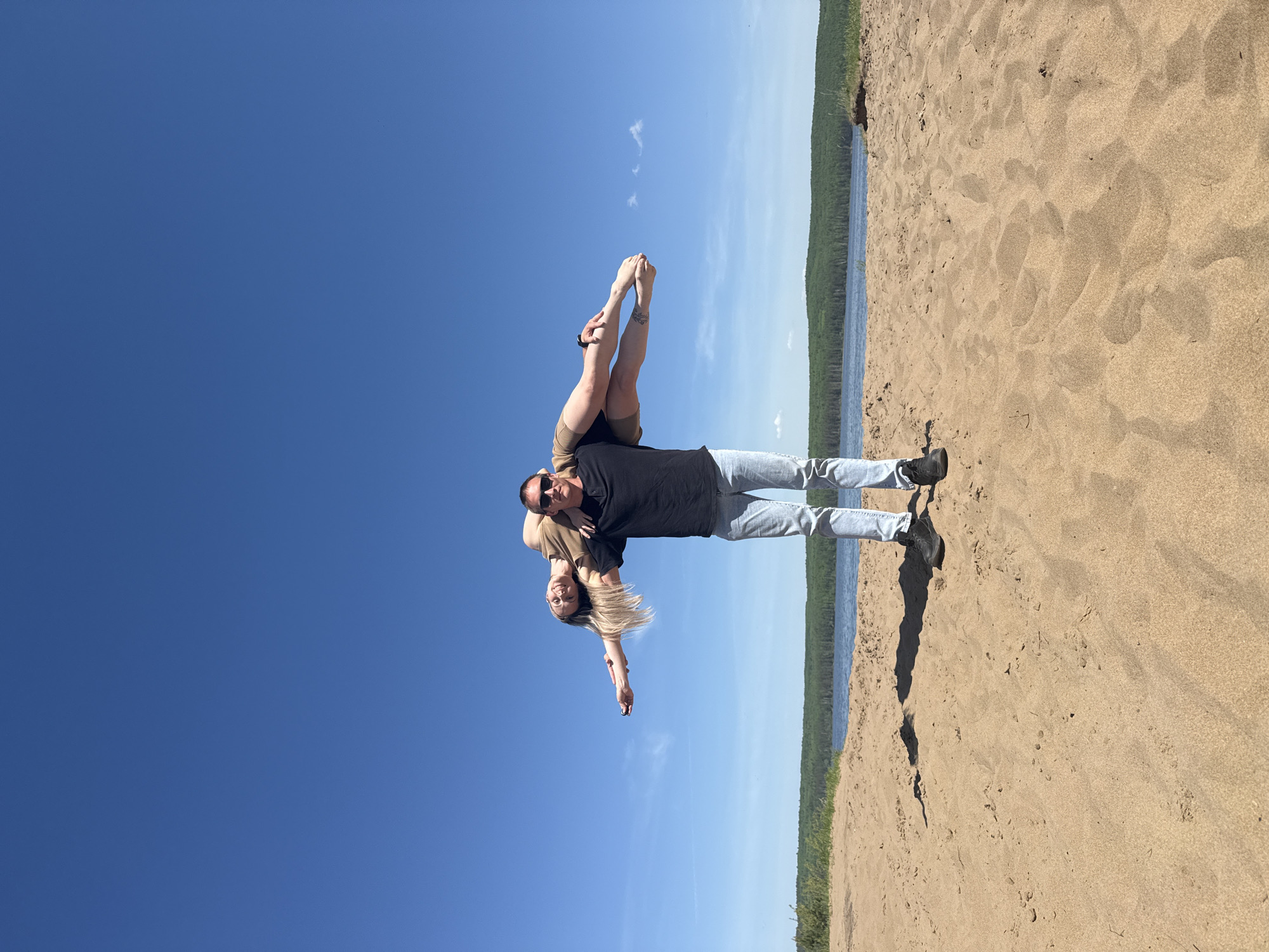 A man and woman are pictured on a sandy beach under a clear, blue sky. The man, wearing a black shirt, light blue jeans, black shoes, and sunglasses, is laying on his back on the beach. The woman, wearing a light-colored top and shorts, is positioned on top of him. Her feet are pointing upward, and she is holding onto her toes. The location appears to be on a beach in Chaikovsky, Russia, based on visual cues in the background showing a body of water and trees.