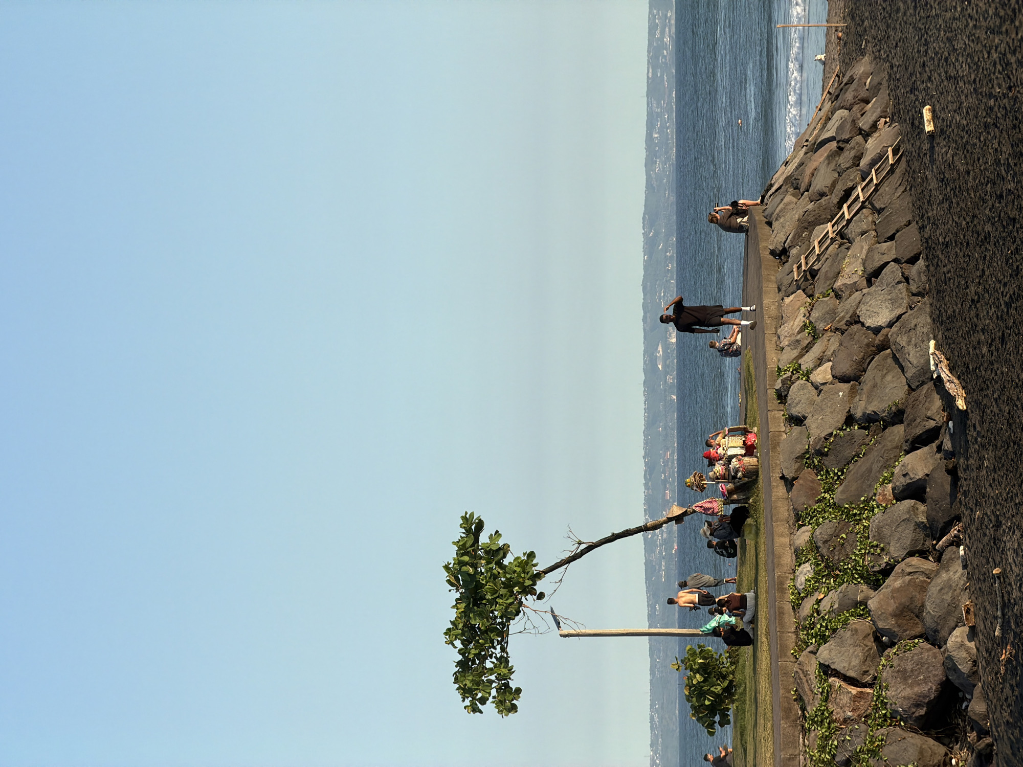 The image depicts a coastal scene in Mengwi, Indonesia. The foreground features a rocky embankment with interspersed vegetation. A leaning tree stands alongside a utility pole. A paved path runs parallel to the embankment.
Several people are present. Some are standing, while others are sitting or reclining. Some individuals appear to be engaged in activities near the water's edge. A ladder leans against the rocks. The background shows the ocean and a distant shoreline with buildings. The sky is clear and light blue. Black sand is also seen in the image.