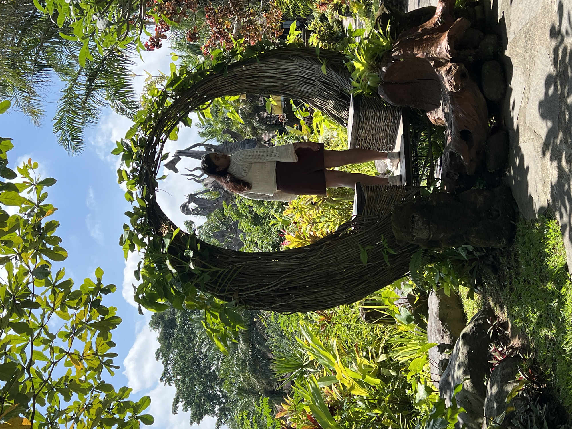A person is standing within a large, circular, woven structure in what appears to be a garden or park setting. The individual is wearing a cream-colored top and a brown skirt. The structure is surrounded by lush green foliage and other plants. The background includes trees and a partly cloudy sky. The location is in Kedewatan, Indonesia.