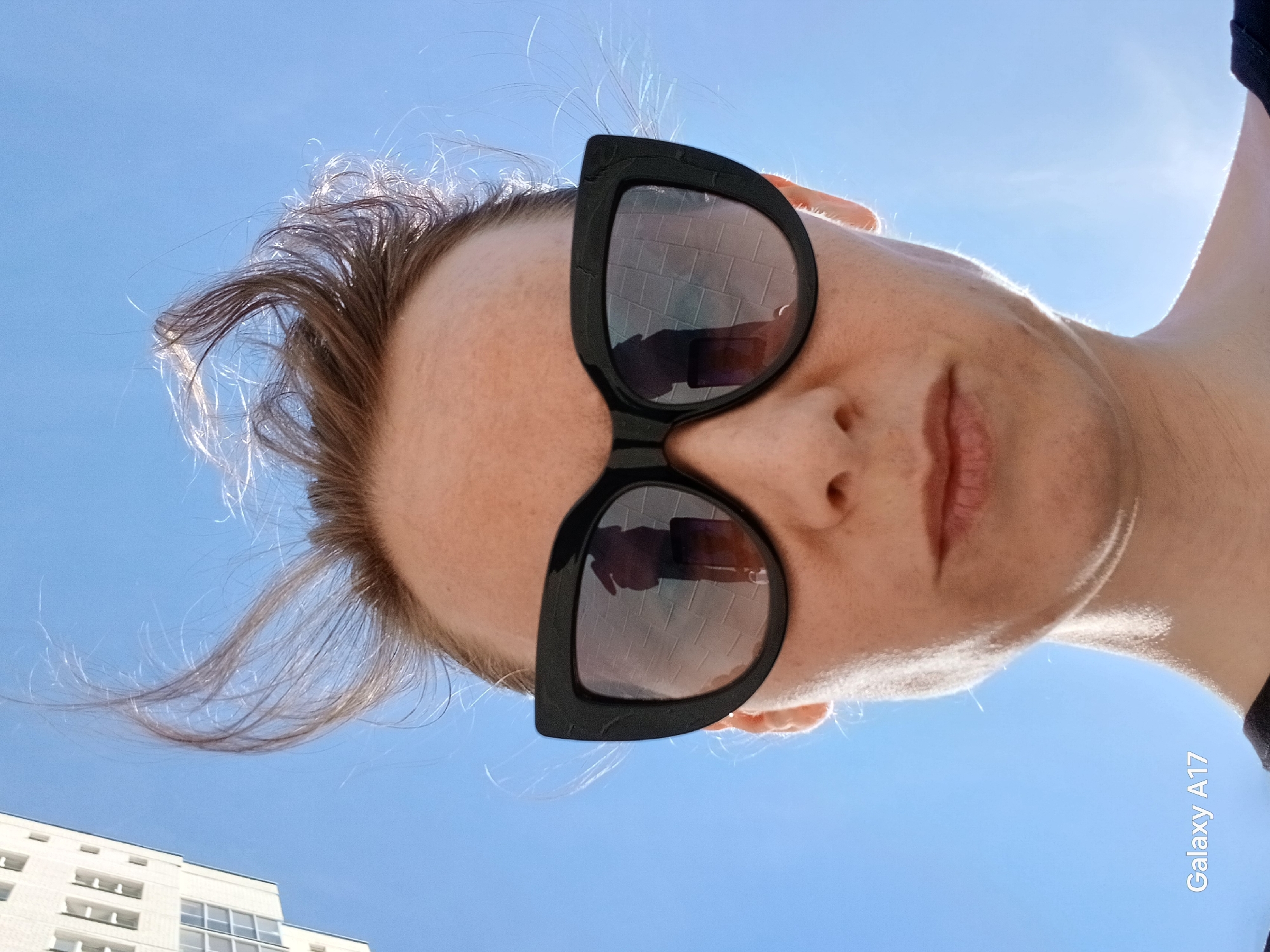 A woman with short brown hair and wearing black sunglasses is looking down at the camera from an upward angle. The sunglasses reflect a building with multiple windows. The sky is blue. In the lower left corner, a partial view of a white building is visible, indicating a possible location in an urban area. The text "Galaxy A17" is on the lower right of the picture indicating the phone used to take the photo. The city of Yekaterinburg, Russia, is not explicitly indicated but can be implied from the reflection in the sunglasses.