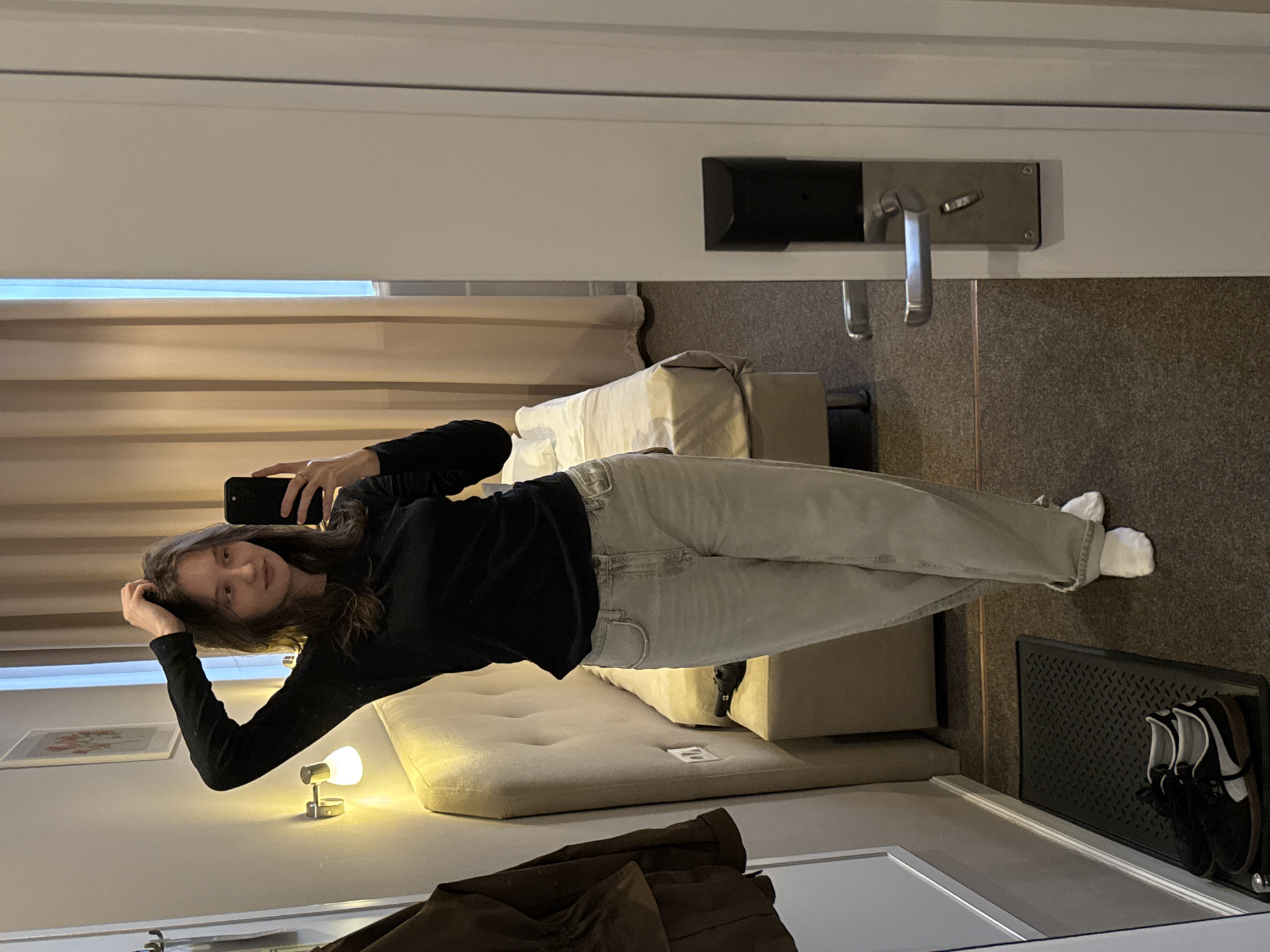 A young woman with brown hair and brown eyes is taking a mirror selfie in a hotel room. She is wearing a black long-sleeved shirt and light-colored jeans. She is standing and has her right hand raised, touching her hair. In her left hand, she is holding a black smartphone, which is pointed at the mirror. The room appears to be a standard hotel room with a bed, a nightstand, and a closet. A lamp is visible on the wall above the headboard of the bed. A door with a silver handle is on the right side of the frame. The floor is carpeted. The overall impression is that of a candid shot taken in a private space. The image is a selfie taken from the user's perspective.