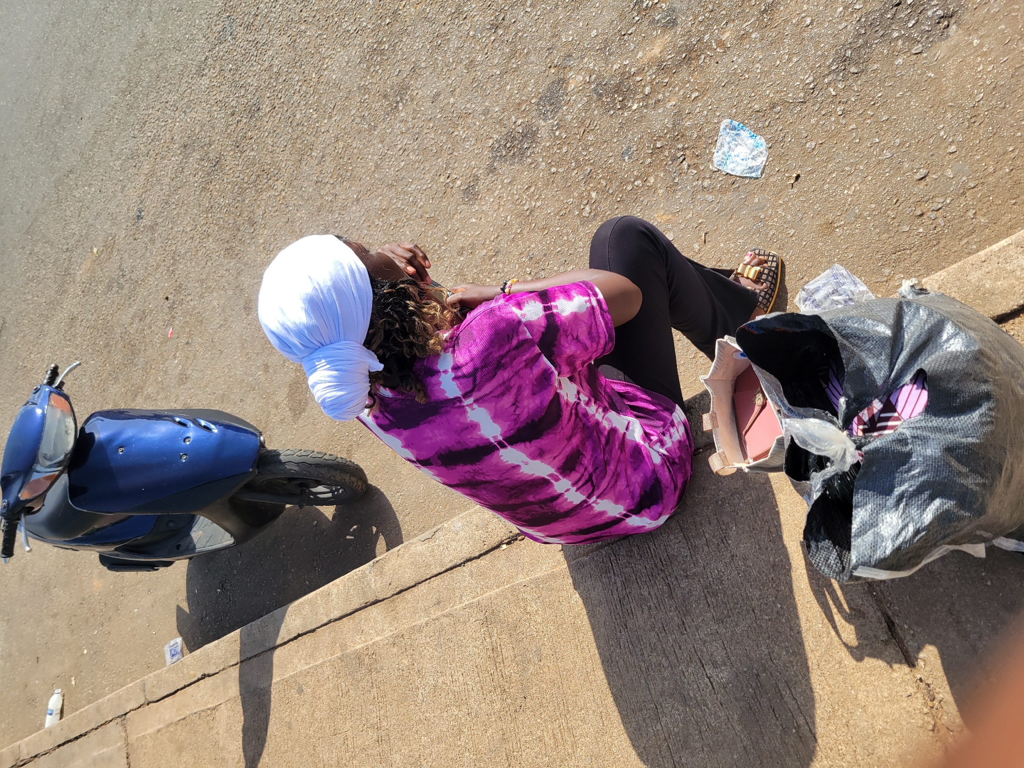 A lone Black person wearing a white headscarf, a pink and white tie-dye t-shirt, and black pants sits on the curb of a paved street, facing away from the camera. The person's right hand is resting on their face. To the left of the person, a blue scooter is parked on the street, its front wheel turned towards the curb. In front of the person, on the curb, is a small, open box with a pink object inside, and a large black plastic bag. Scattered on the street are pieces of trash. The scene appears to be in the city of Jos, Nigeria.