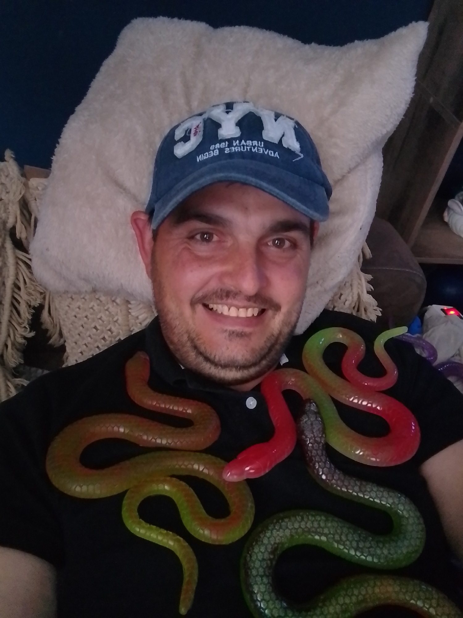 A man wearing a blue baseball cap that says "NYC" is lying down, with his head resting on a large fluffy pillow. He is smiling at the camera. Several colorful rubber snakes are draped around his neck and chest, over a black collared shirt. The snakes are red, green, and iridescent grey. The background shows a bookshelf with various objects, suggesting an indoor setting. The specific location in the city of Wedel, Germany, is not discernible from the image.