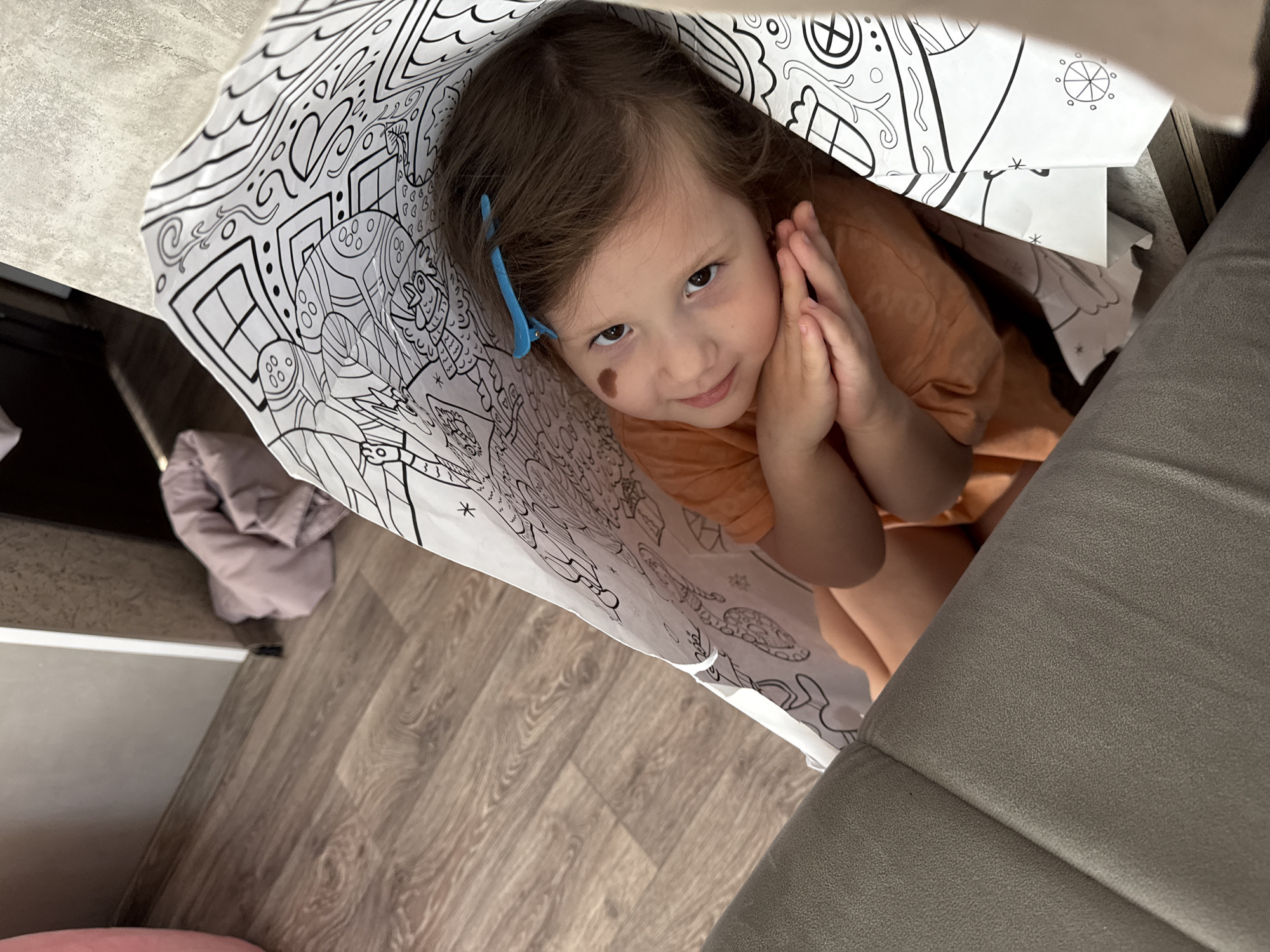 A young girl with brown hair and a blue clip is nestled inside a large, white, patterned paper bag. Her hands are clasped together, and she is looking directly at the camera with a slight smile. She has a visible birthmark on her left cheek. The bag appears to be filled with coloring book style illustrations. The bag is resting on a light-colored wooden floor and a grey upholstered sofa is visible on the right side of the frame. A neutral-colored jacket or blanket is partially visible on the left. The location, Кипчак, Russia, cannot be determined from the image.
