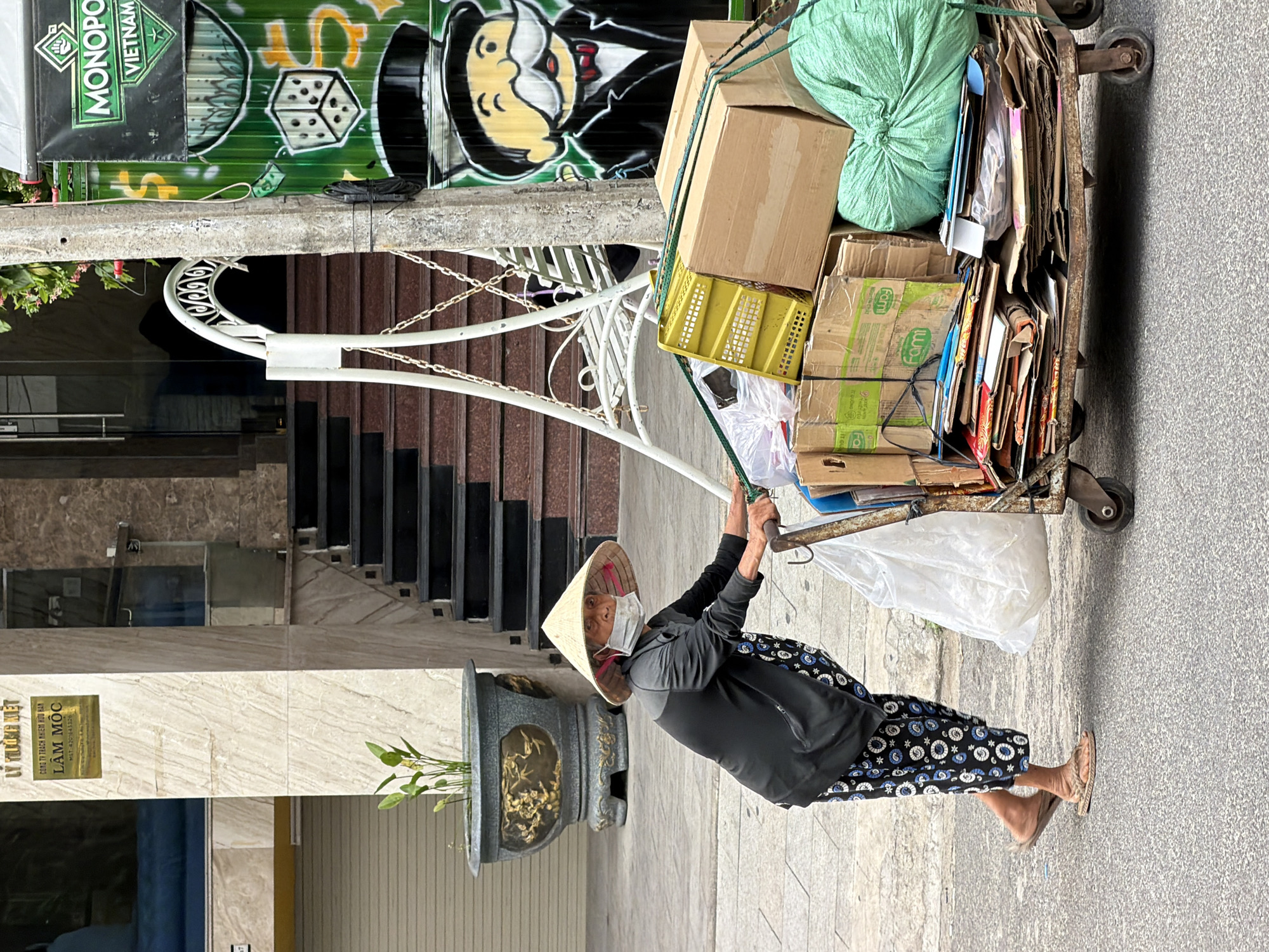 A woman wearing a conical hat and face mask pushes a loaded cart on a sidewalk in Nha Trang, Vietnam. The cart is filled with cardboard boxes, a large green bag, and other assorted items. A building with a sign that reads "LÂM MỘC" is visible in the background, along with a large planter containing a plant. A pole with graffiti related to Monopoly Vietnam stands nearby. The woman is dressed in a dark grey long-sleeved shirt and patterned pants, and she wears simple flip-flops. The overall scene depicts a common urban activity of waste collection or delivery in a Vietnamese city.