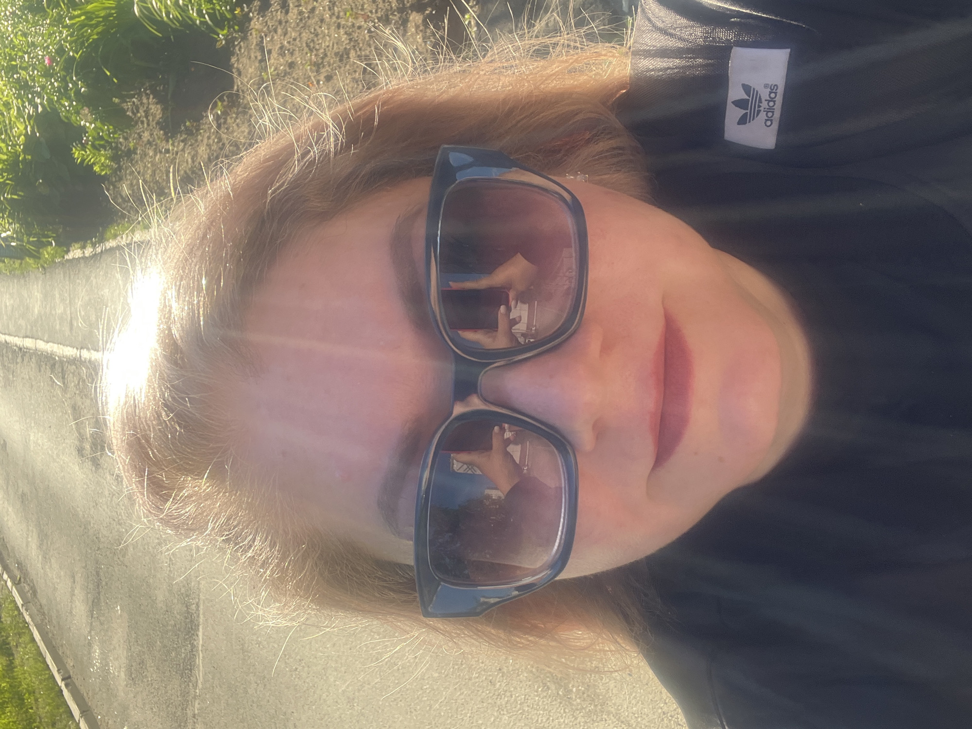 A woman is taking a selfie outdoors. She is wearing large, dark-rimmed sunglasses and a black t-shirt with an Adidas logo on the sleeve. Her blonde hair is slightly windblown. The background shows a paved path and a grassy area with sunlight filtering through. The scene appears to be in a park or along a roadside in Izhevsk, Russia, based on the provided context. The reflection in her sunglasses shows her holding a phone and taking the picture.