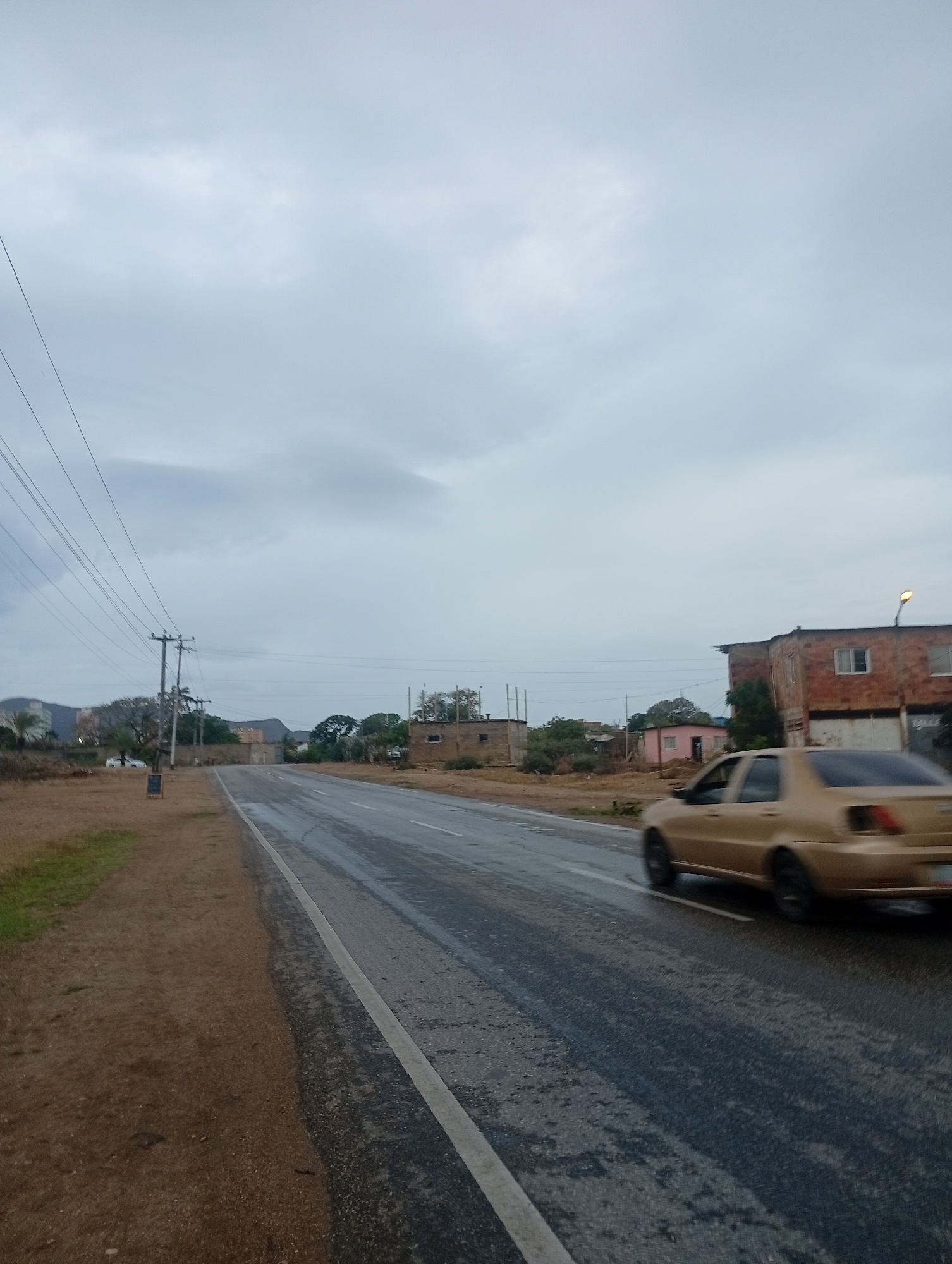 The image depicts a rural road scene in Juan Griego, Venezuela, under an overcast sky. A two-lane asphalt road, wet from recent rain, curves gently to the left. A tan sedan is captured in motion on the right lane, its image blurred due to speed. On the left side of the road, a dirt shoulder is visible, with sparse dry grass. Several power lines run parallel to the road, supported by wooden utility poles. In the background, various structures are scattered along the roadside, including low-rise brick buildings, some appearing dilapidated, and a small pink building. Trees and vegetation are present between the buildings. In the far distance, mountains are faintly visible. A signpost stands on the left shoulder near a utility pole. The overall atmosphere is subdued, likely due to the cloudy weather.