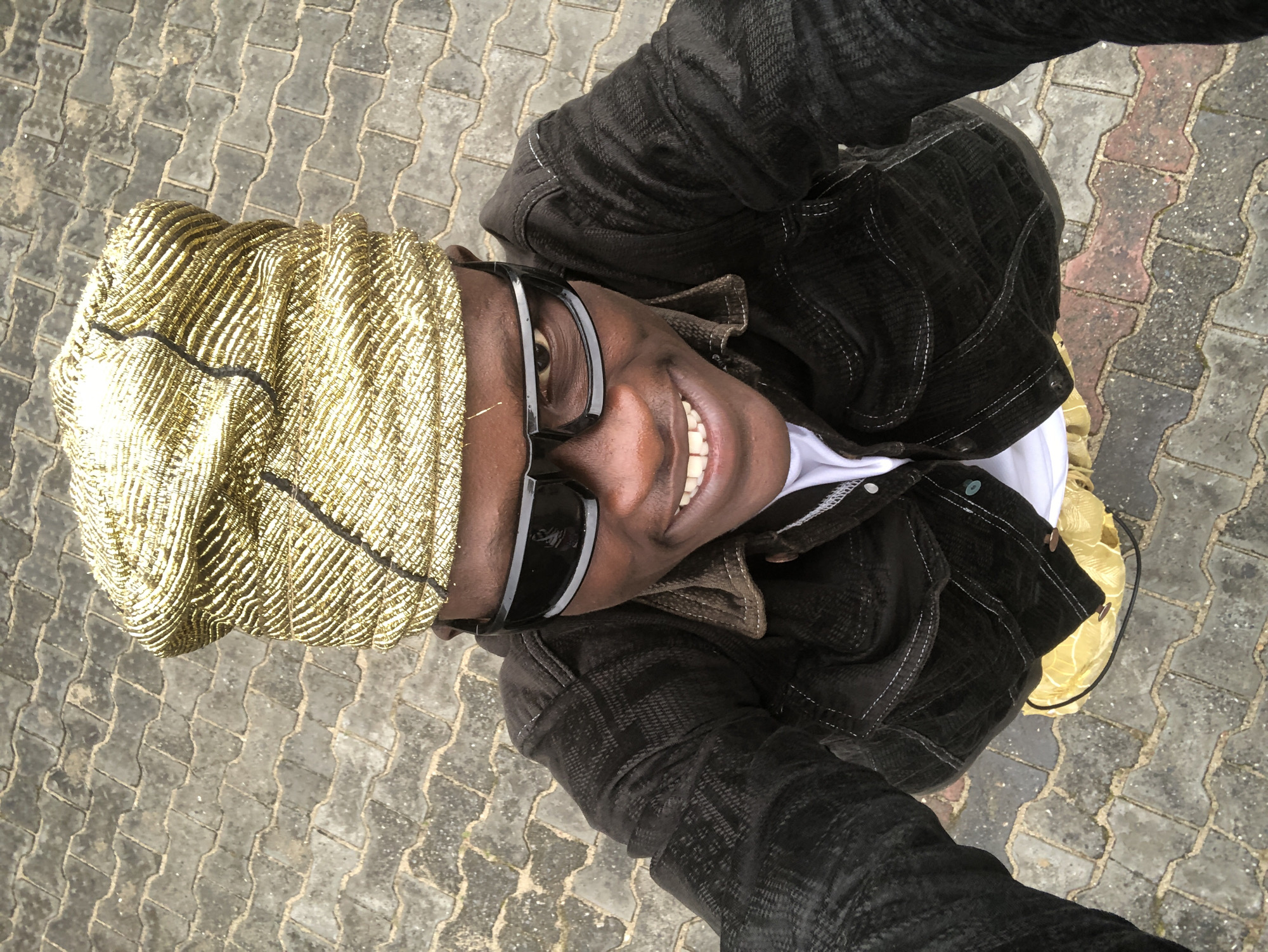 A high-angle, full-body selfie shows a young Black male standing on a paved outdoor area in Ado, Nigeria. He is smiling and wearing a gold, crinkled headwrap, black sunglasses with thick frames, a dark, textured button-down jacket over a white t-shirt, and shiny yellow shoes. The ground beneath him is paved with interlocking concrete pavers. The image appears to have been taken during daylight.