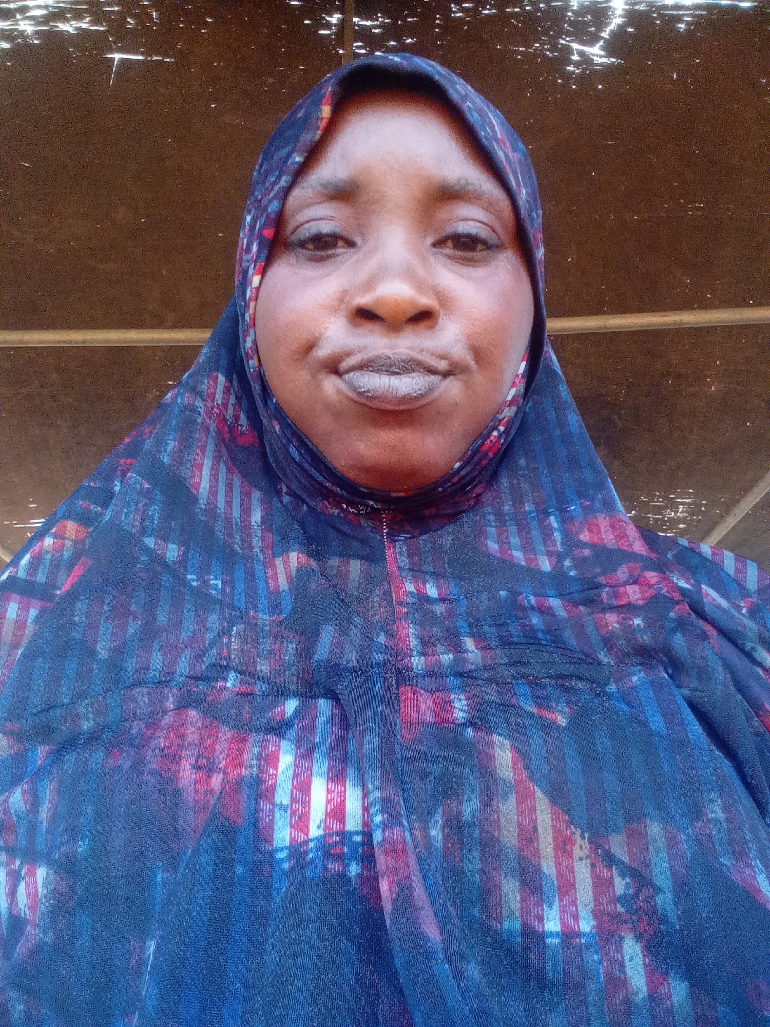 A medium eye-level close-up shot depicts a woman wearing a patterned hijab. She appears to be located in an area in Akwanga, Nigeria. Her face is slightly wrinkled, and she has full lips. The hijab features vertical stripes in shades of blue, red, and white. The background shows a simple wooden structure.