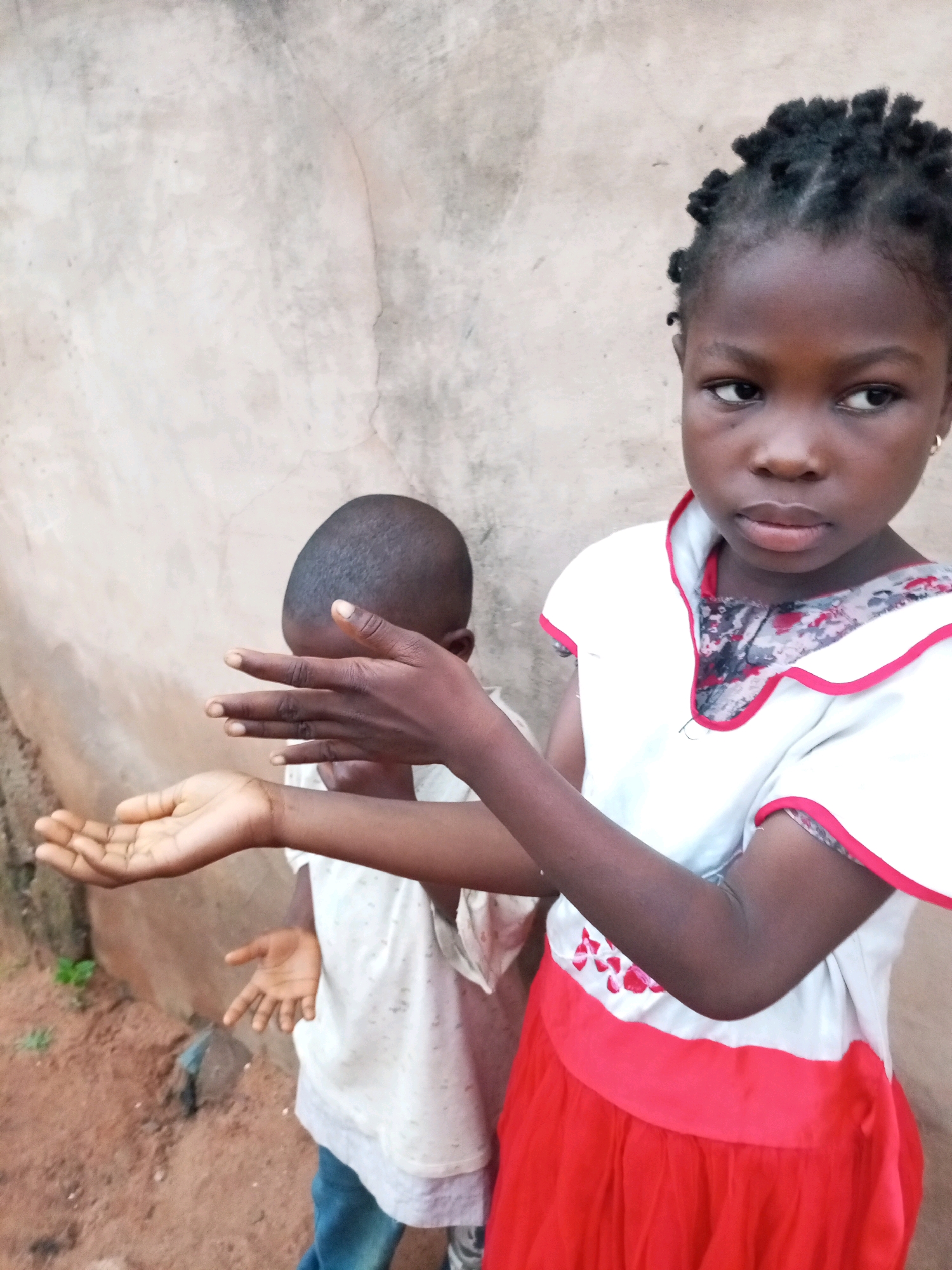 In Tempure, Nigeria, two young children stand against a textured, neutral-colored wall. The child in the foreground, a girl, faces slightly away from the viewer, her gaze directed to her right. She has dark skin and her hair is styled in small, braided twists. She wears a white dress with red trim around the collar and sleeves, and a bright red skirt. The second child, positioned behind and to the left of the girl, is largely obscured from view. This child is also dark-skinned and appears to be wearing a white t-shirt. Their head is turned away, and their hands are raised in front of their face, with one hand held palm-up in a gesture of offering or receiving. The ground beneath them is reddish-brown earth, with some sparse green vegetation visible to the left. The overall scene depicts a candid moment between two children.